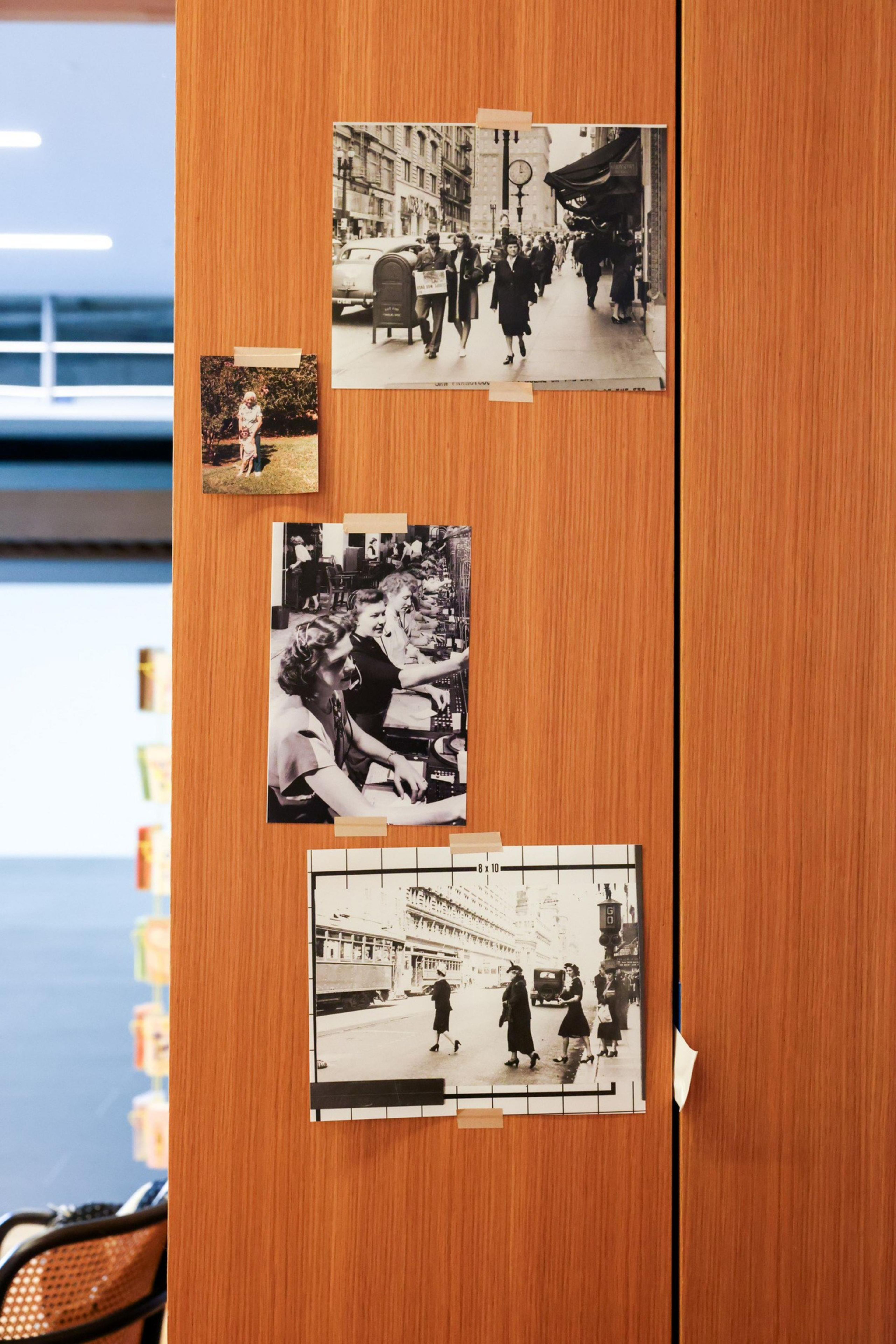 Black-and-white photos are taped to a wooden surface: a city street scene, women working at desks, pedestrians crossing a street, and a small outdoor snapshot.
