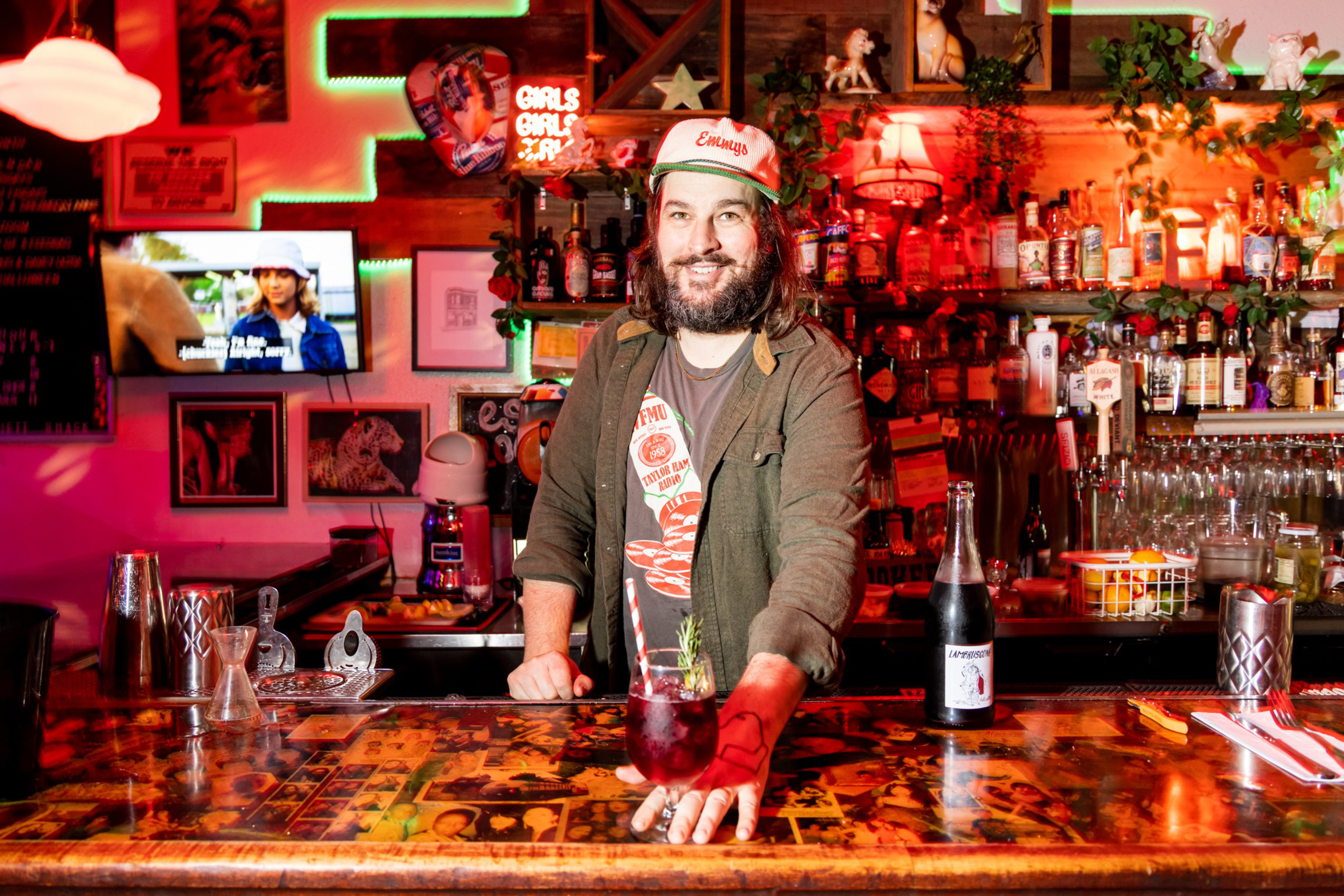 A bearded man wearing a cap stands behind a bar with a red drink in front of him, surrounded by bottles and colorful neon lights.