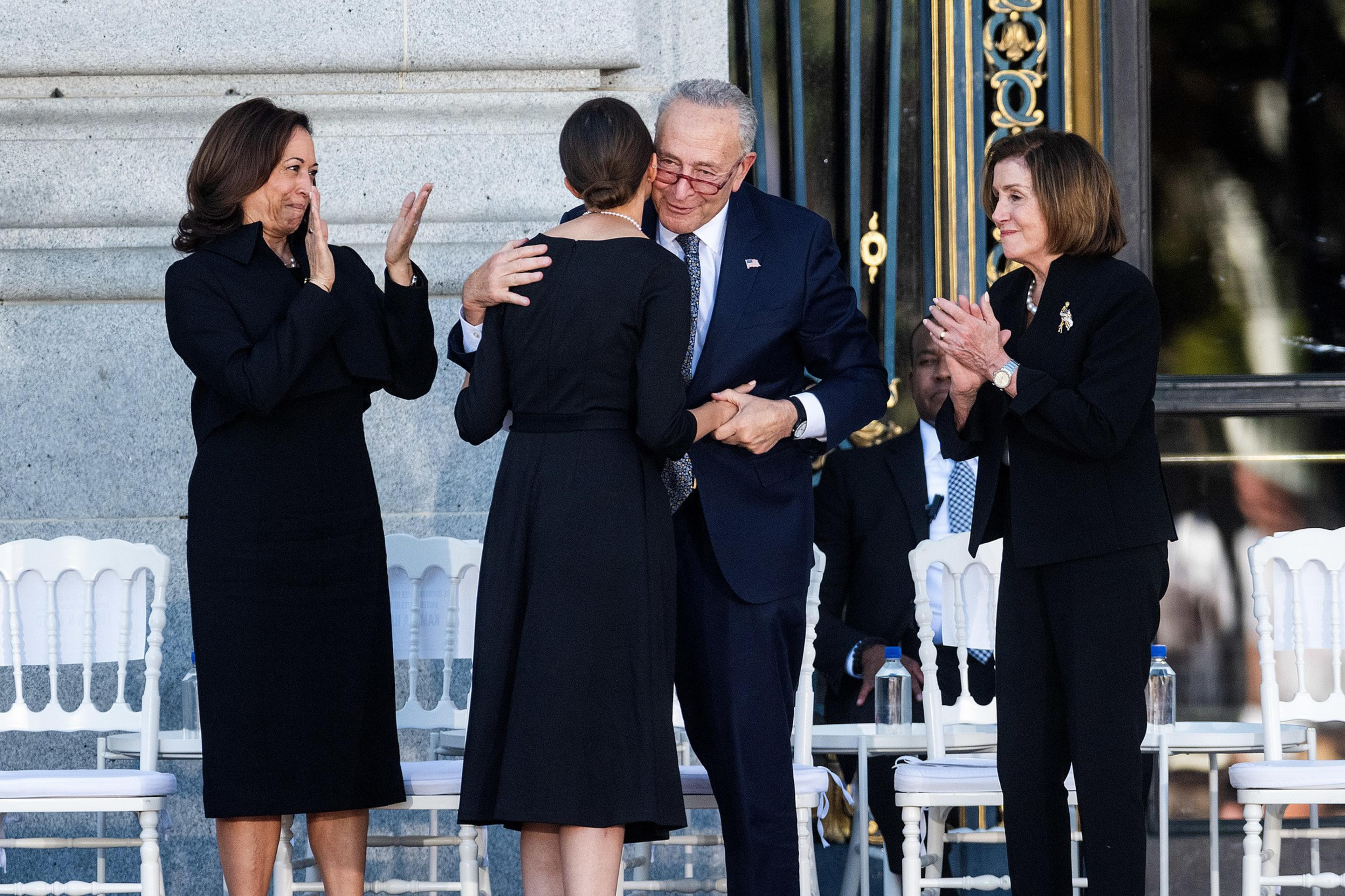 Four people stand in formal attire. Two are facing each other, embracing and shaking hands. The others are clapping, with chairs and a large doorway behind them.