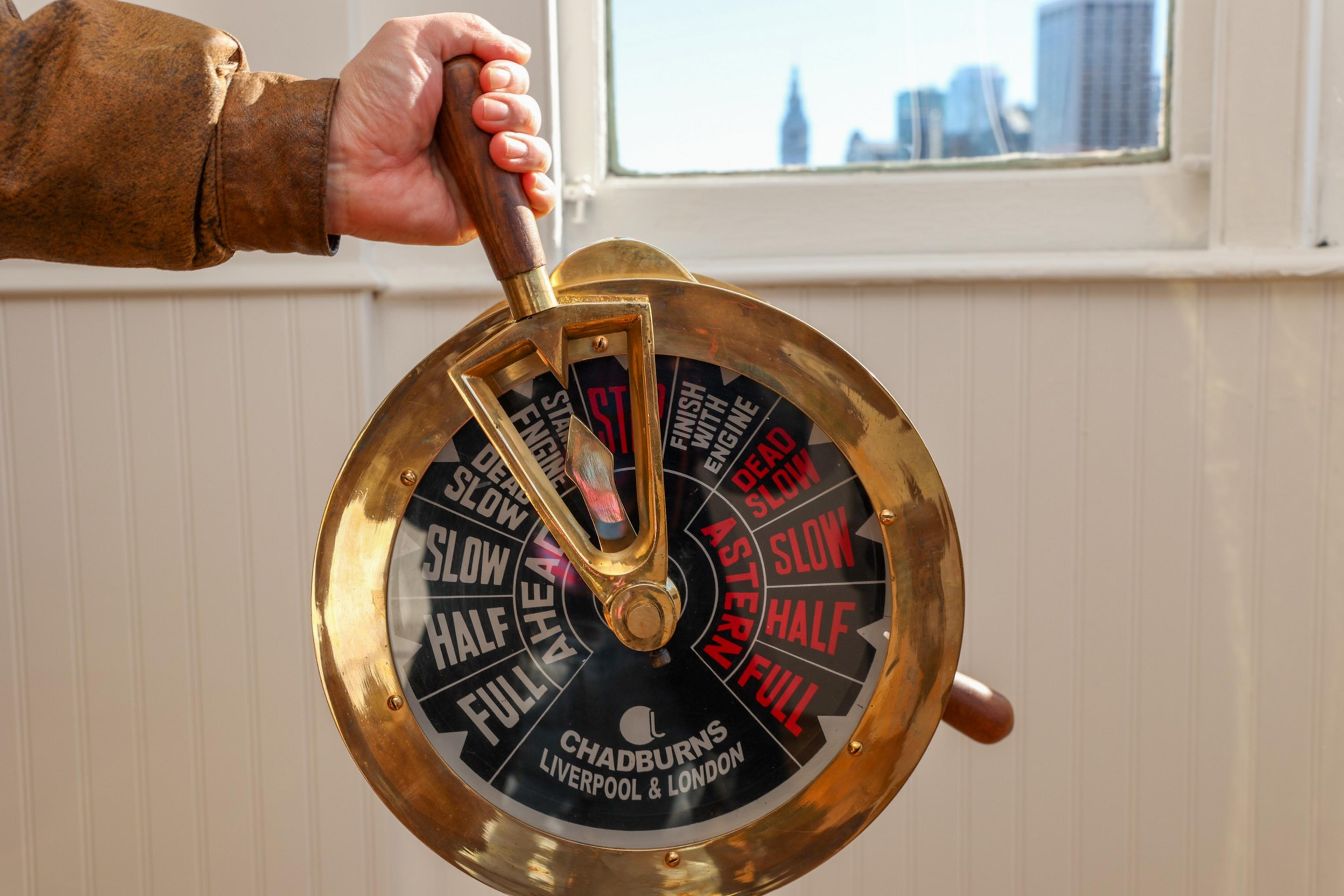 A hand adjusts a vintage brass ship engine telegraph with settings ranging from "Full Ahead" to "Full Astern," labeled "Chadburns Liverpool & London."