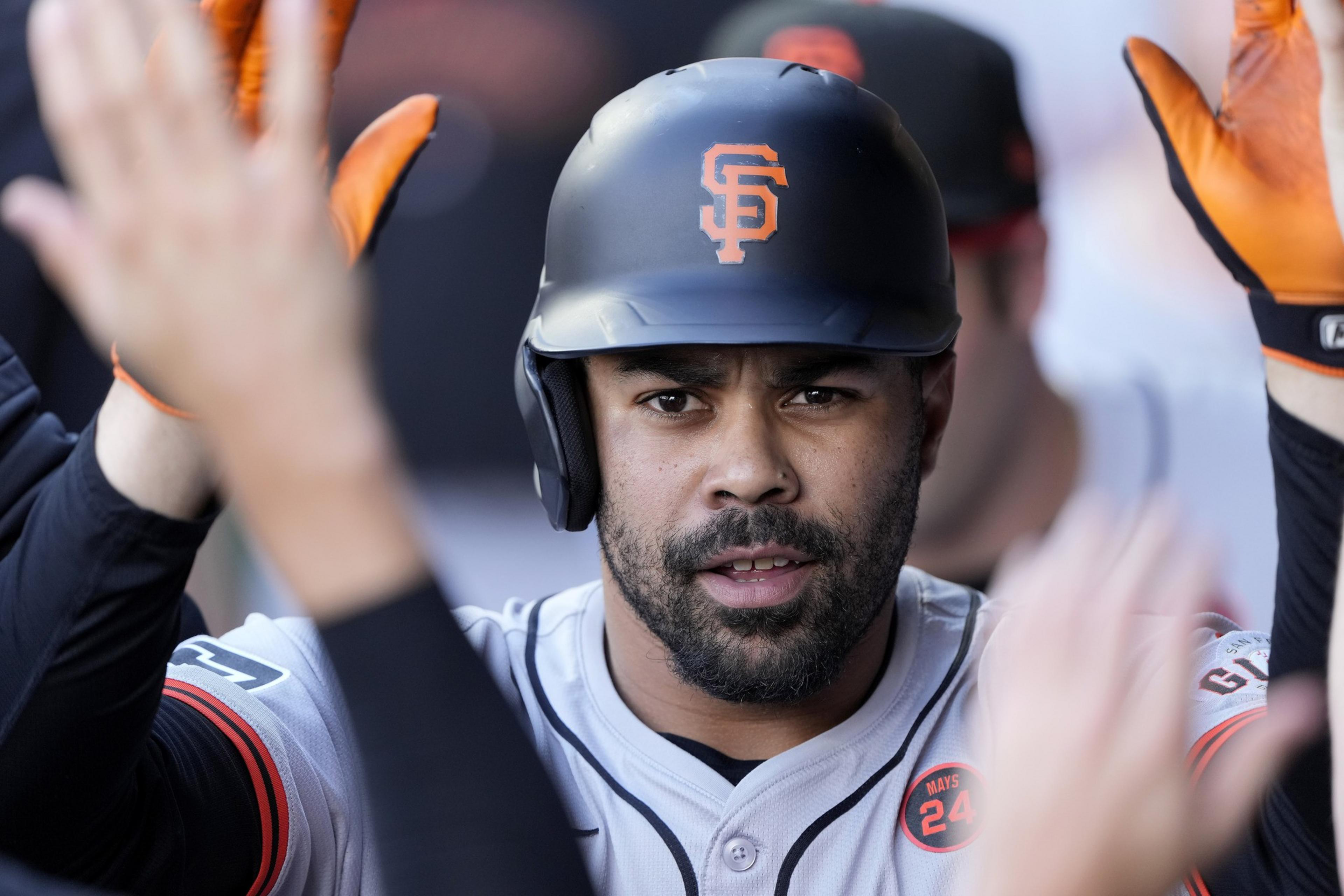 A baseball player wearing a black helmet with an orange logo is giving high-fives while wearing orange gloves. He's in a gray jersey with button details.