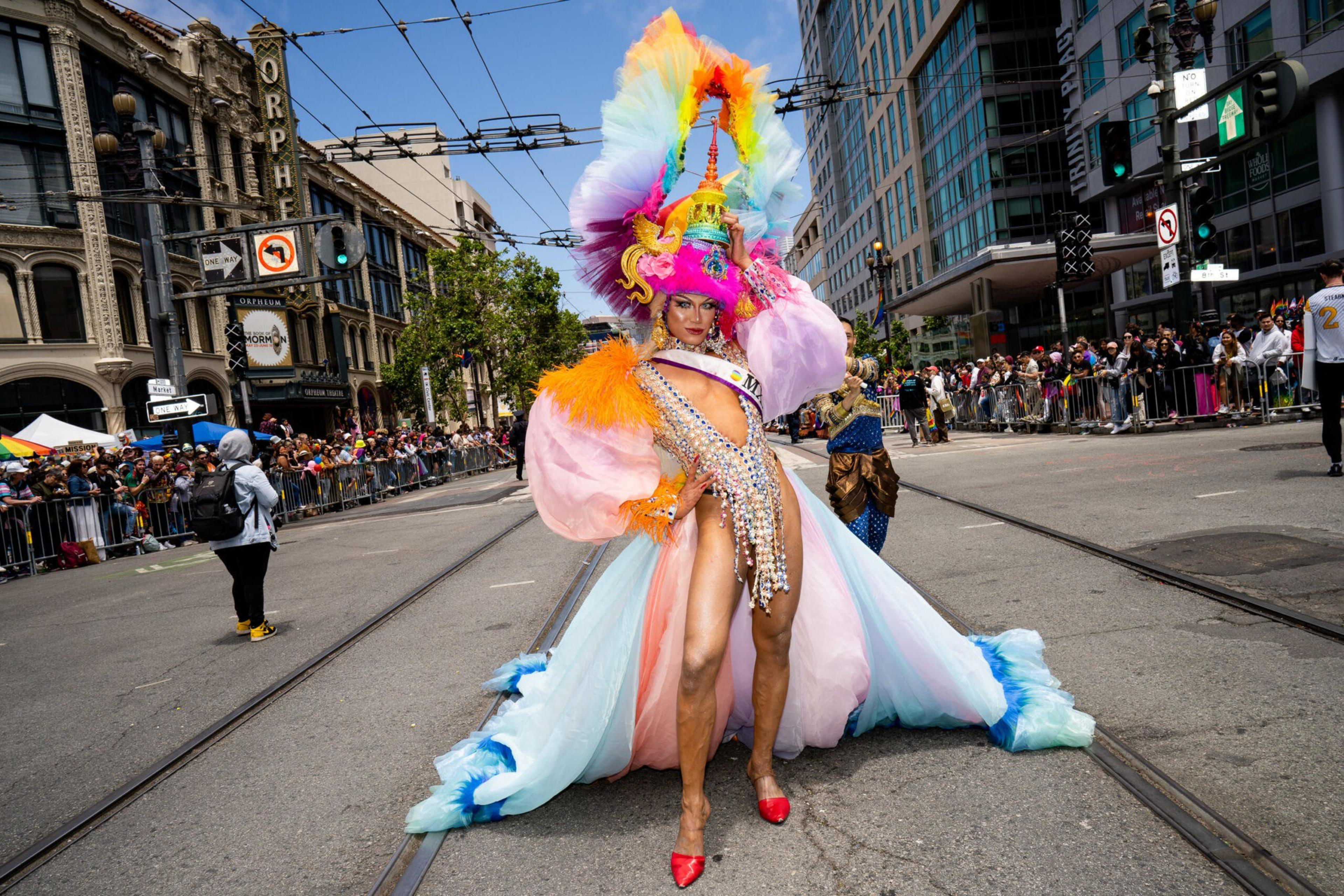 A person in a vibrant, elaborate costume with a tall, multicolored feathered headpiece and flowing pastel train poses confidently on a city street during a parade.