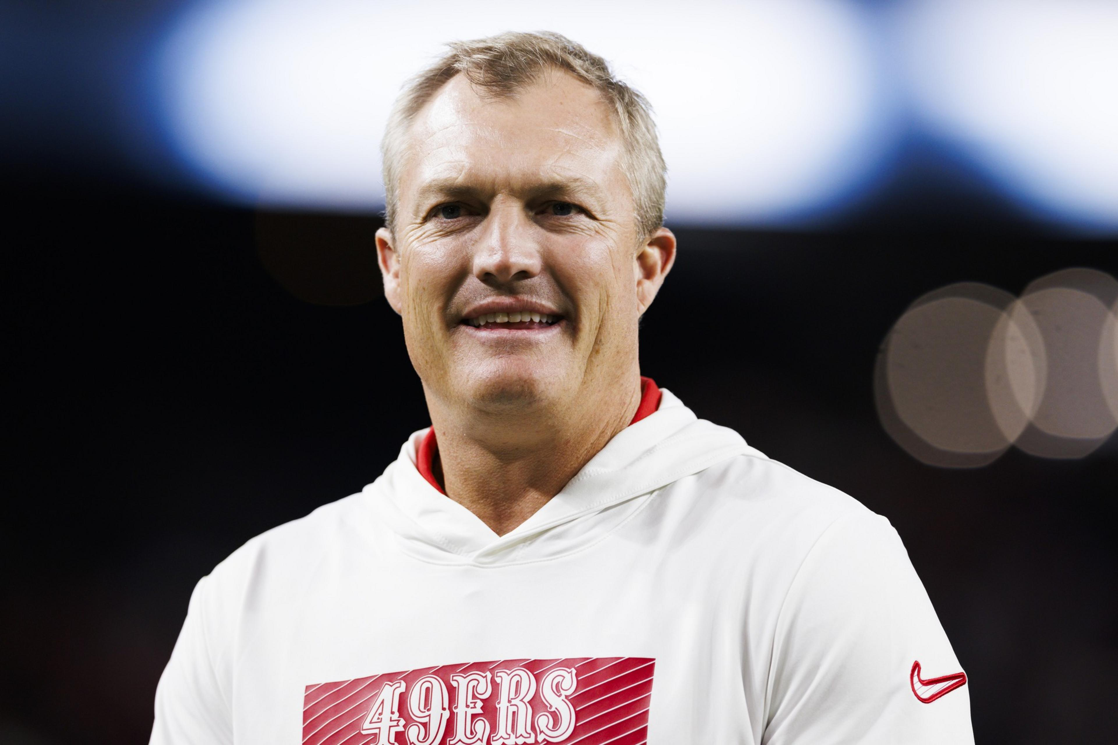 A smiling man is wearing a white hoodie featuring a red panel with "49ERS" on it. The background is blurred, with bright, out-of-focus lights.