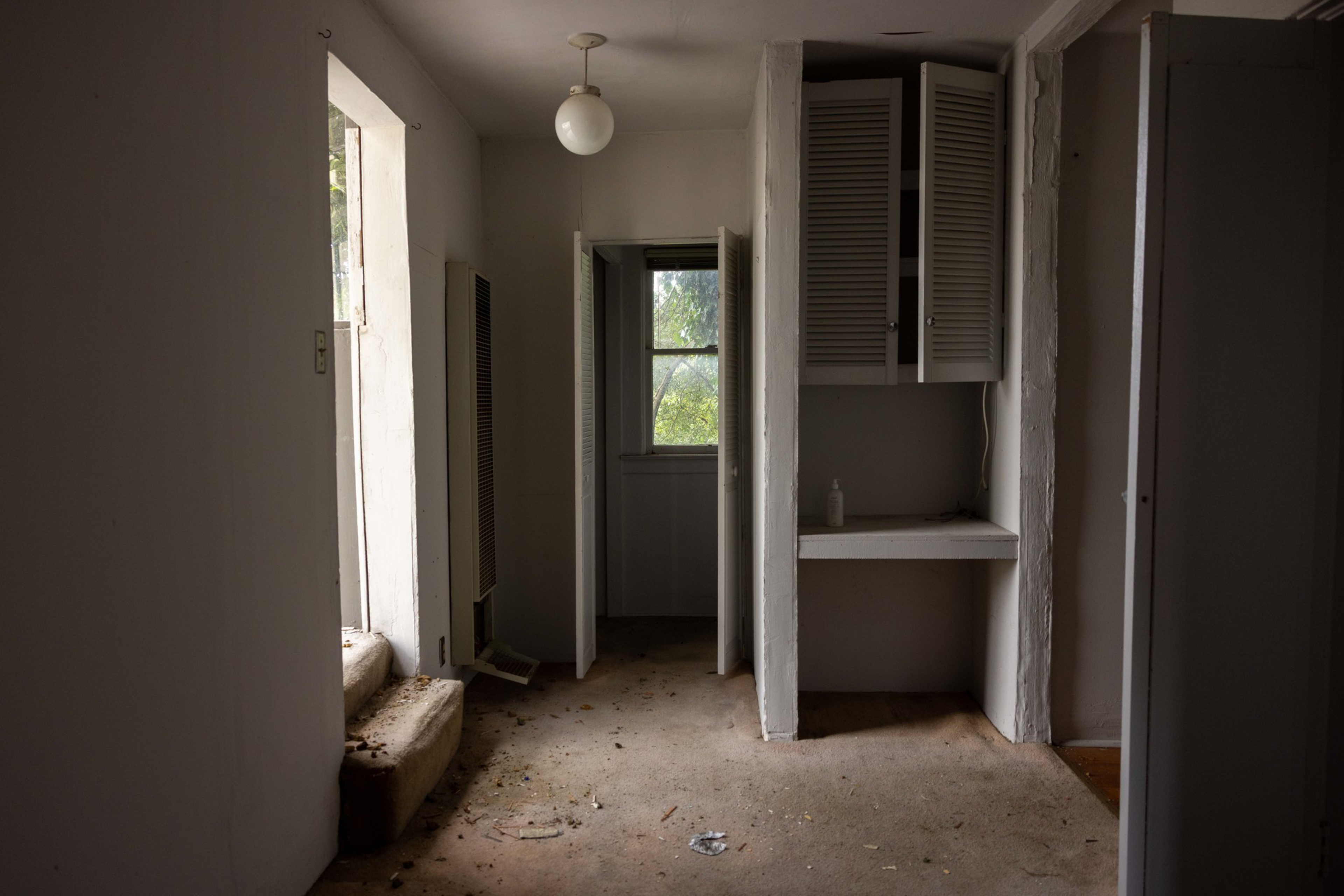 An empty, dimly lit room with dirty carpet, a small built-in desk with shuttered cabinets above, and an open closet door revealing a window.