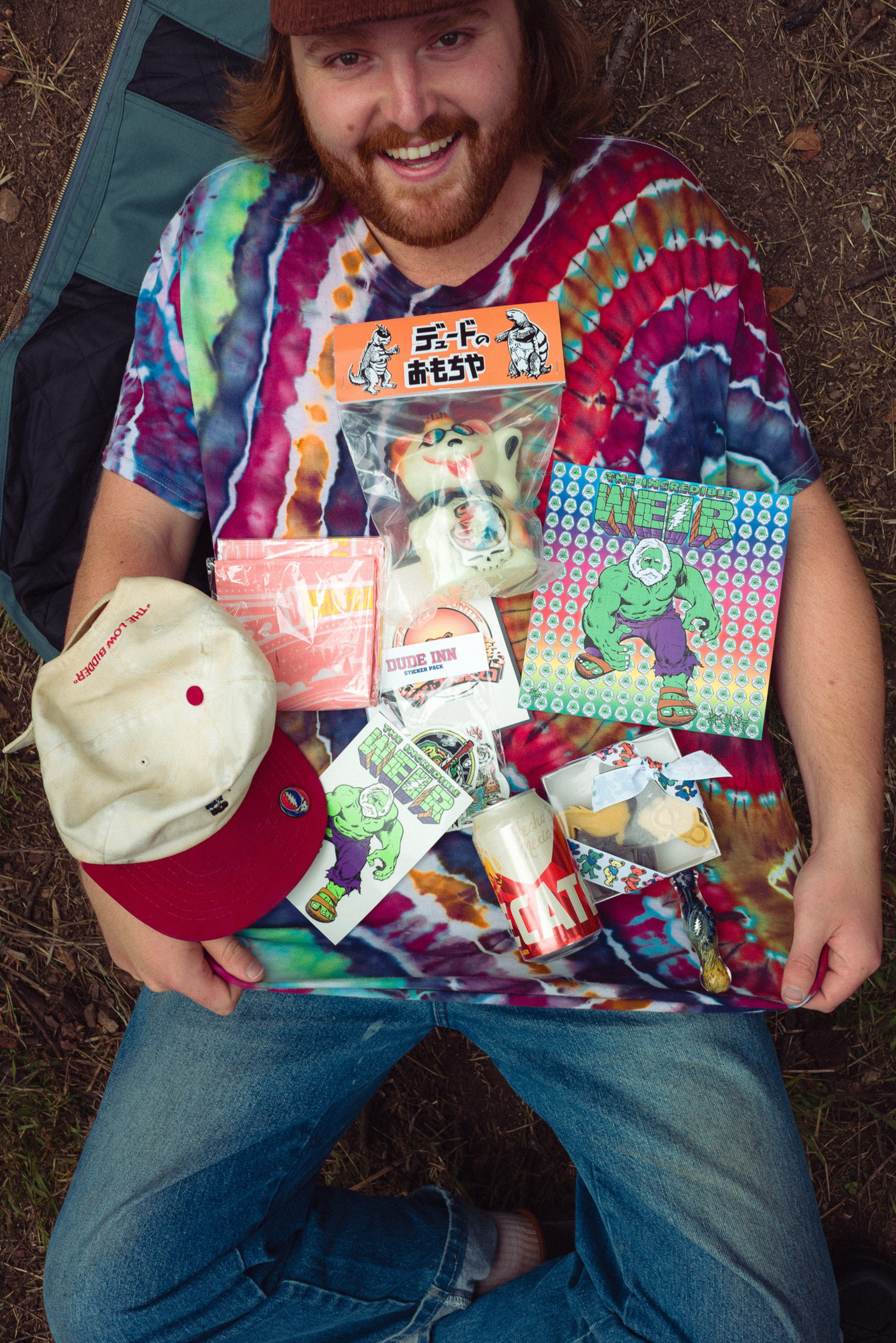 A smiling person wearing a tie-dye shirt and jeans holds various colorful items including a white-red cap, stickers, a toy in packaging, and a can on their lap.