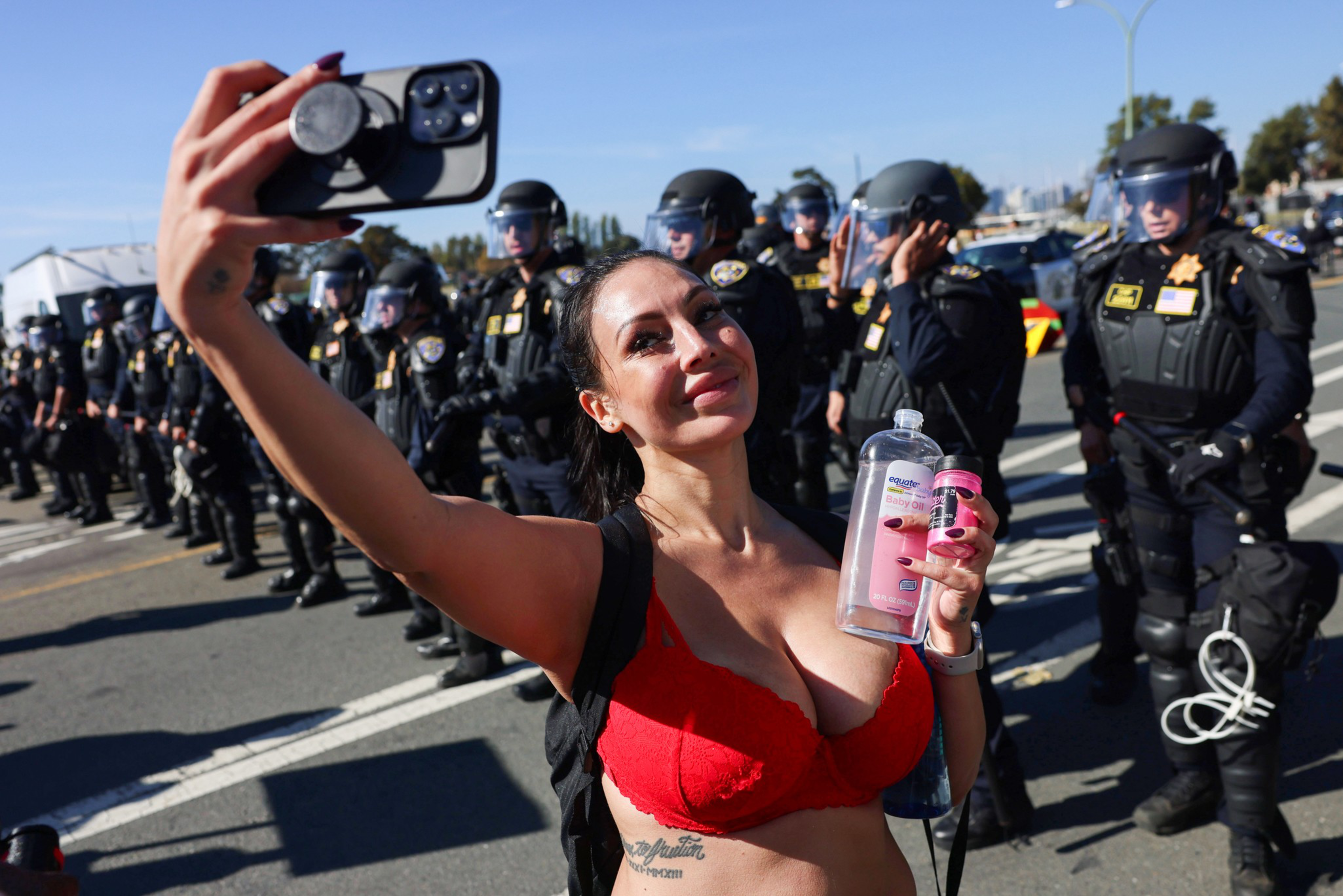 A woman in a red bra takes a selfie holding a water bottle and pink can, while a line of police officers in riot gear stands behind her.