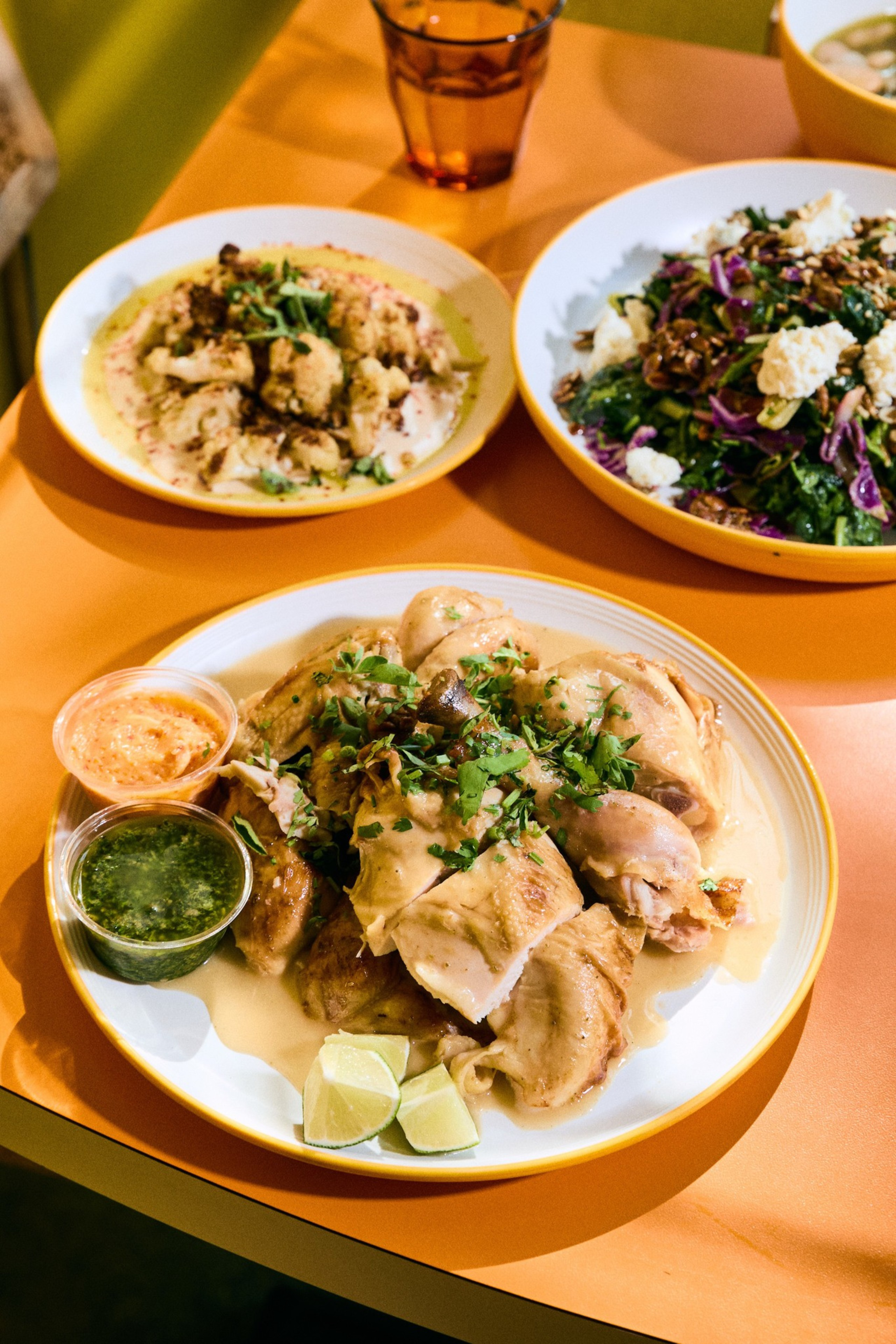 A plate of roasted chicken with herbs, lime wedges, and two dipping sauces is foregrounded. In the background are bowls of cauliflower and a mixed salad.