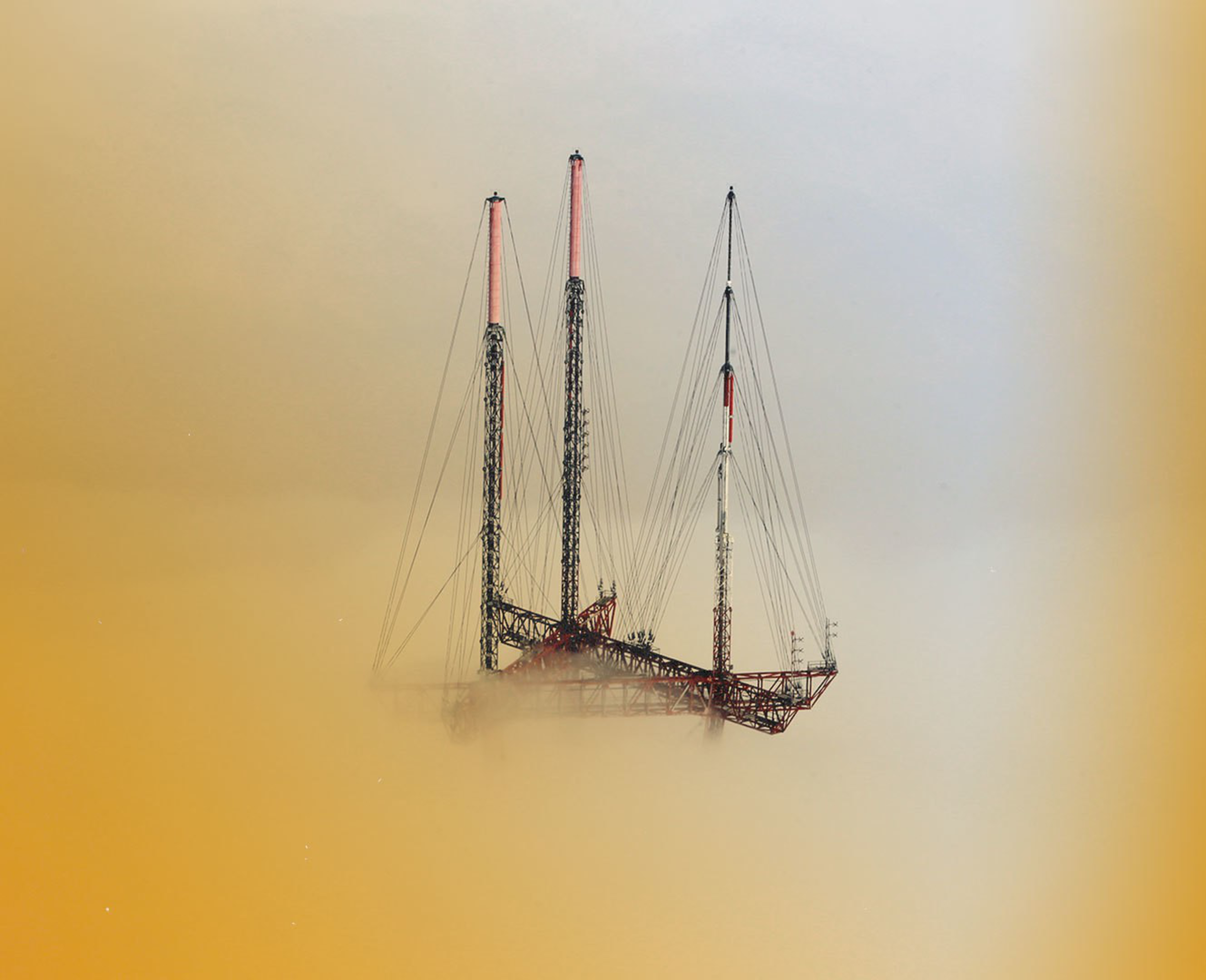 Three tall metal communication towers with cables are partially obscured by orange-brown fog or smoke, blending into a hazy, muted background.