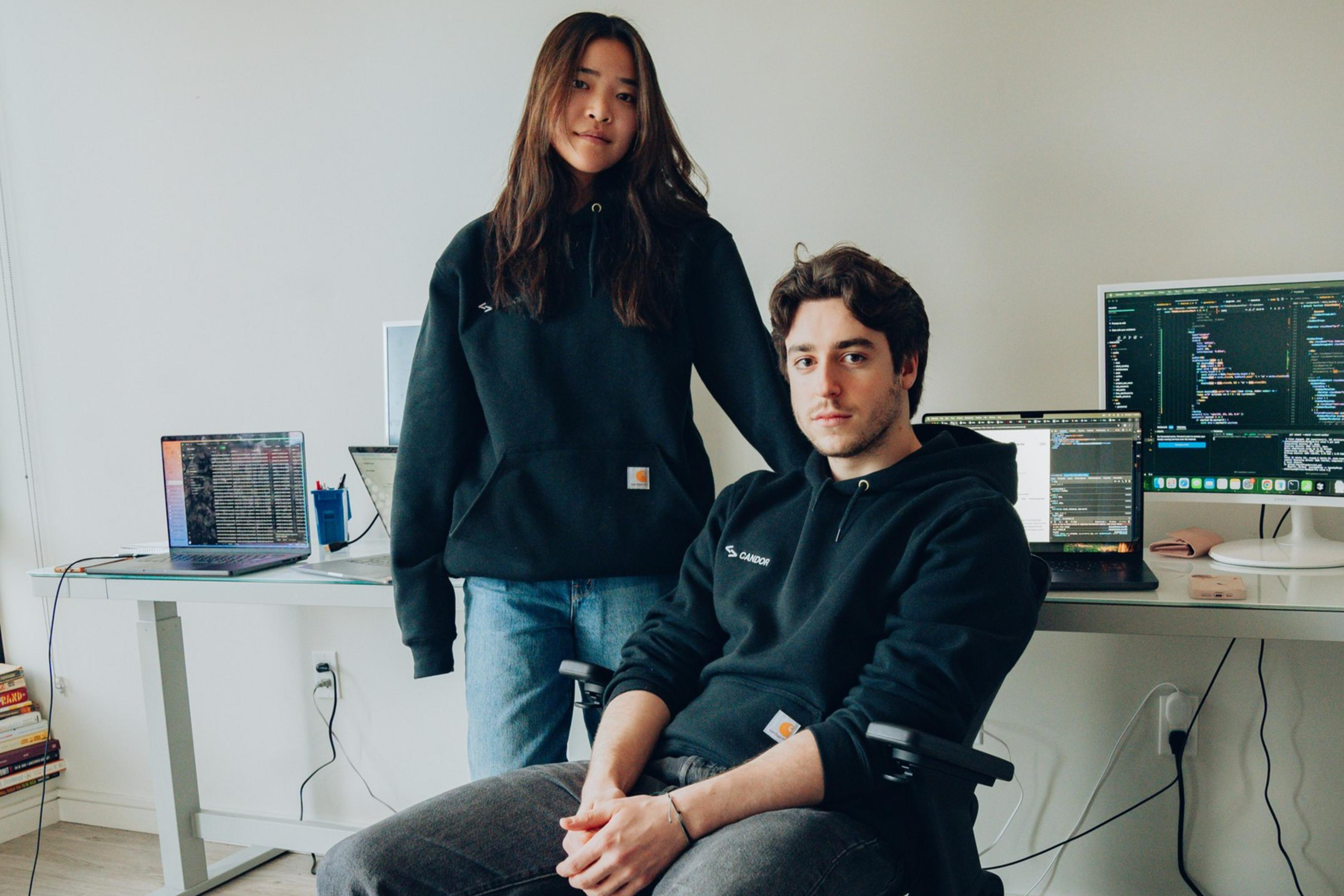 A young woman stands next to a seated man in a home office with computers and code on screens. Both wear black hoodies, and the room is minimalistic and tidy.