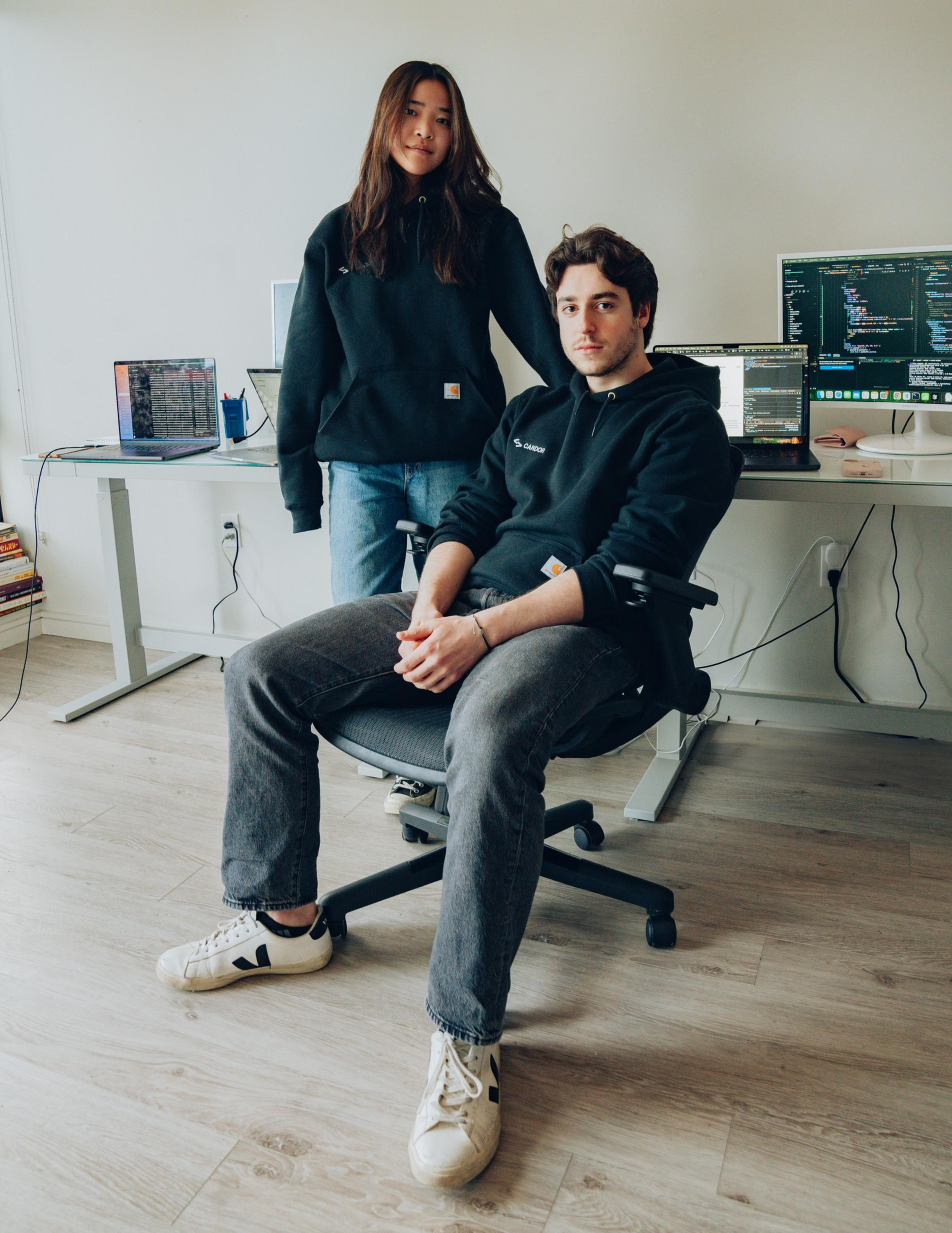 A young woman stands next to a seated man in a home office with computers and code on screens. Both wear black hoodies, and the room is minimalistic and tidy.