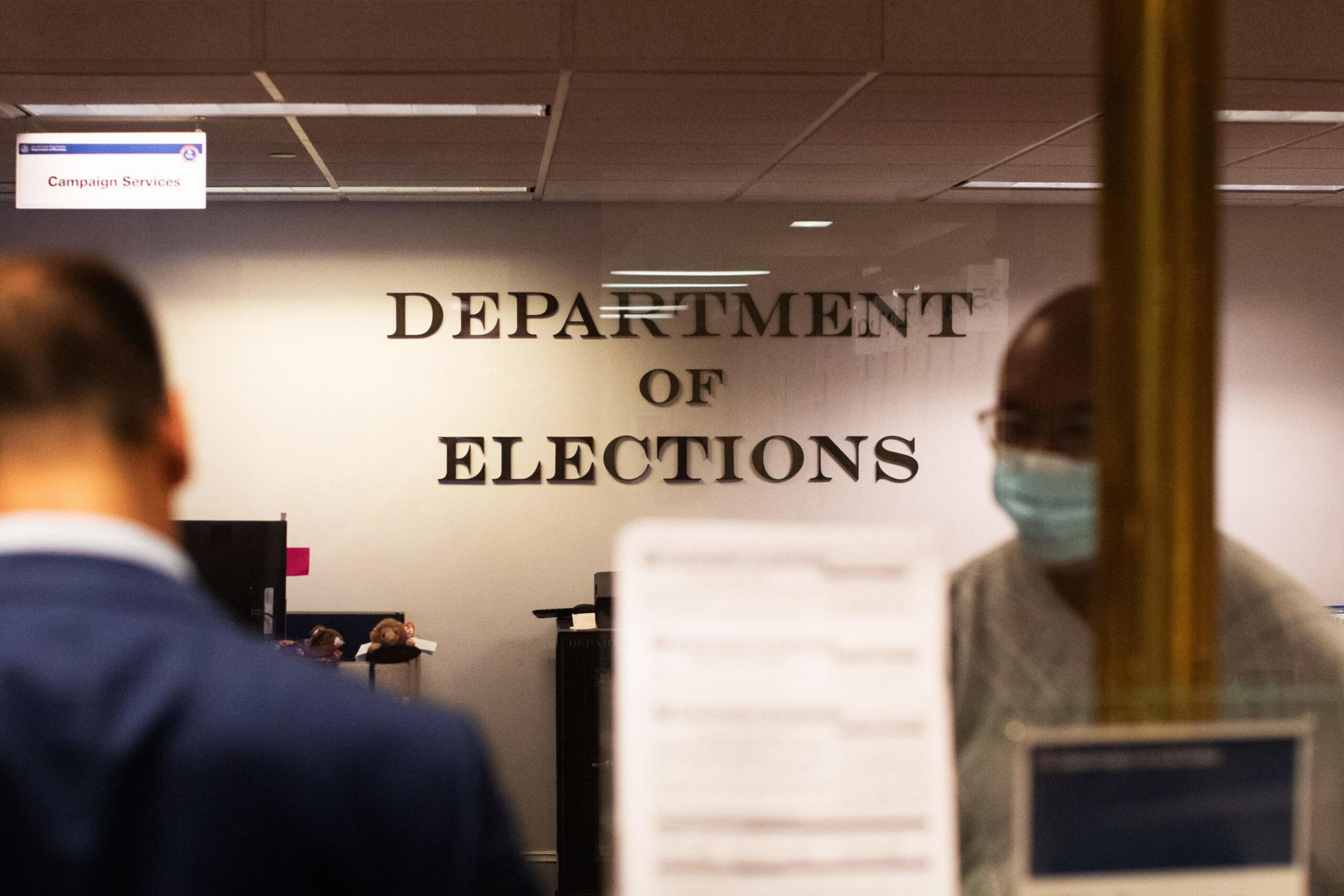 An office with &quot;DEPARTMENT OF ELECTIONS&quot; on the wall, blurred figures in the foreground, possibly through a window.