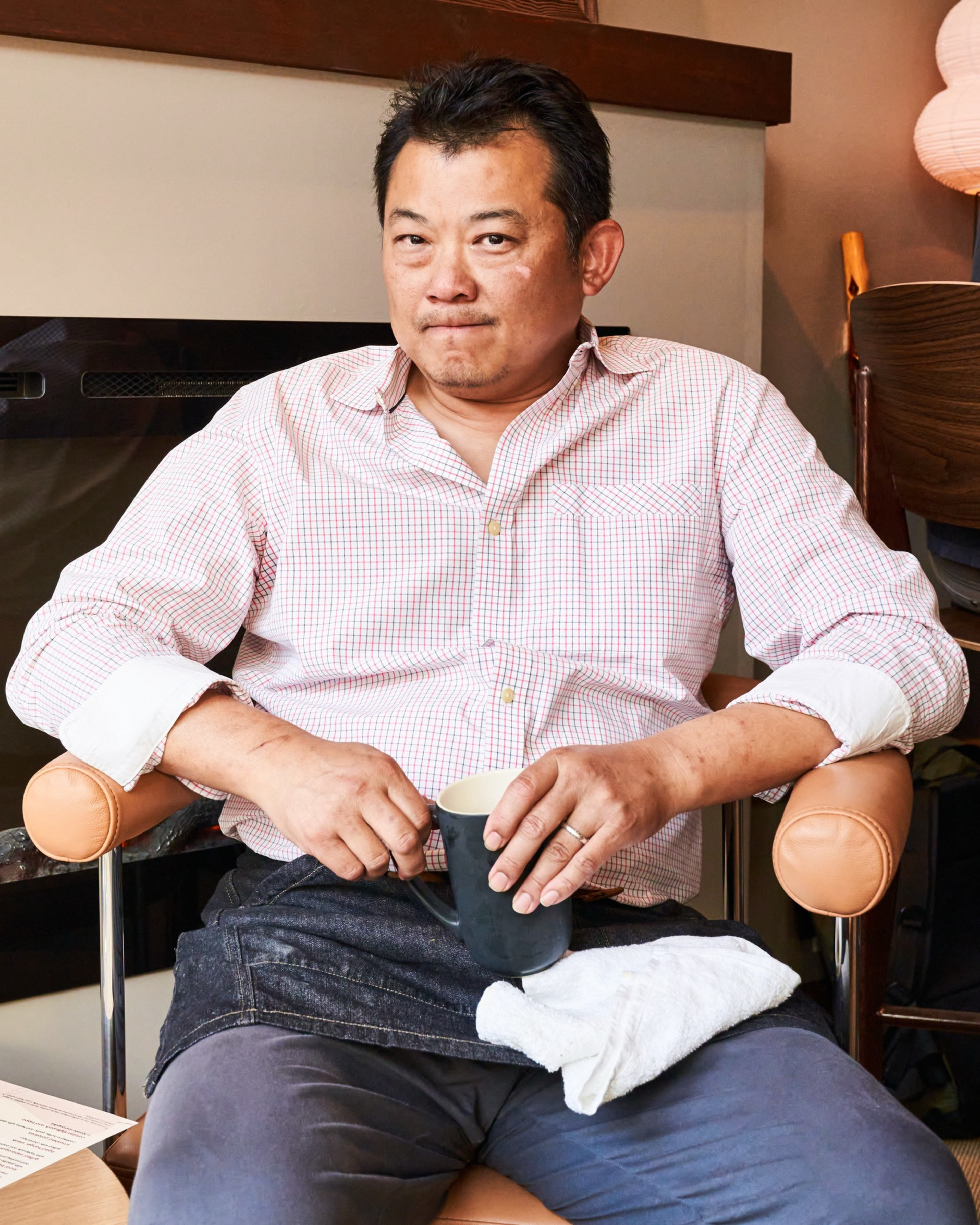 A man in a checkered shirt sits on a chair, holding a black mug and a white towel on his lap. The background has a wooden shelf and a paper lantern.