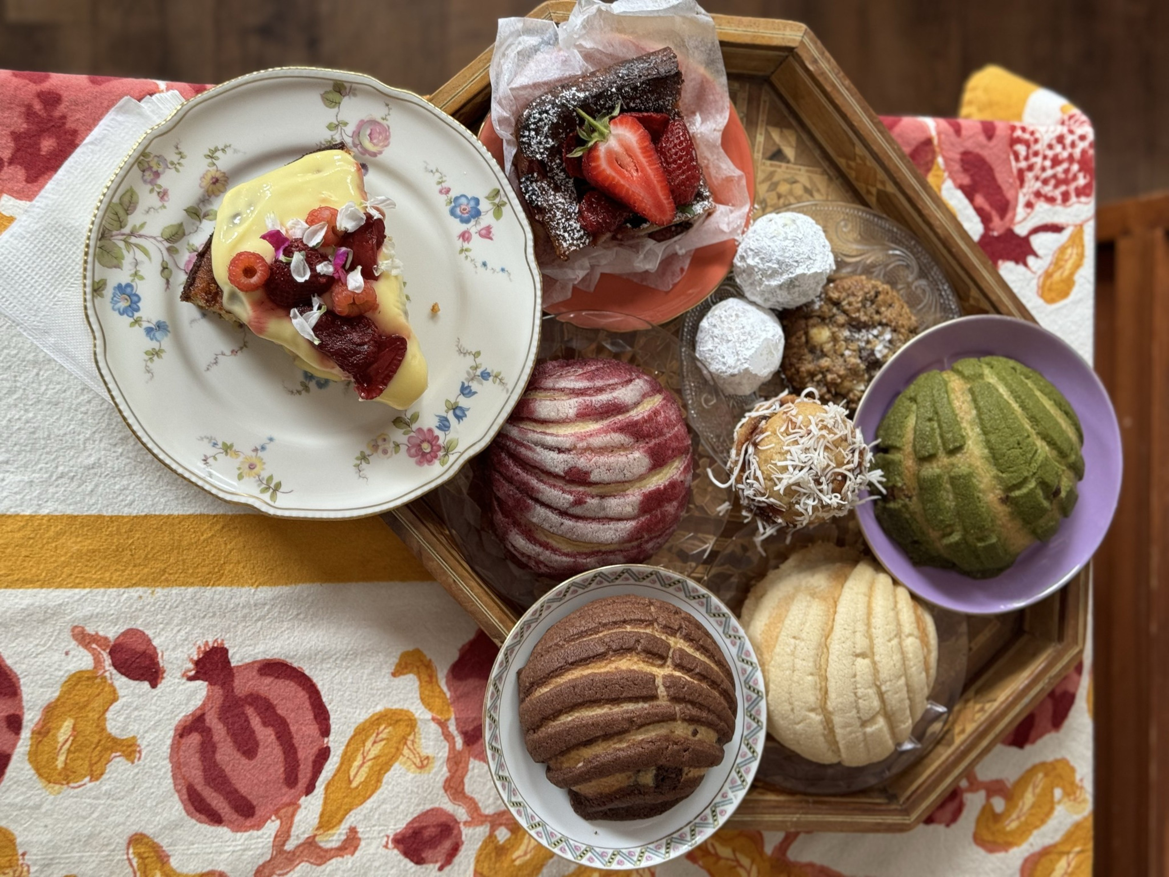 A wooden tray holds colorful conchas, powdered cookies, and a chocolate brownie topped with strawberries, while a floral plate displays a slice of cake with cream and fruit.