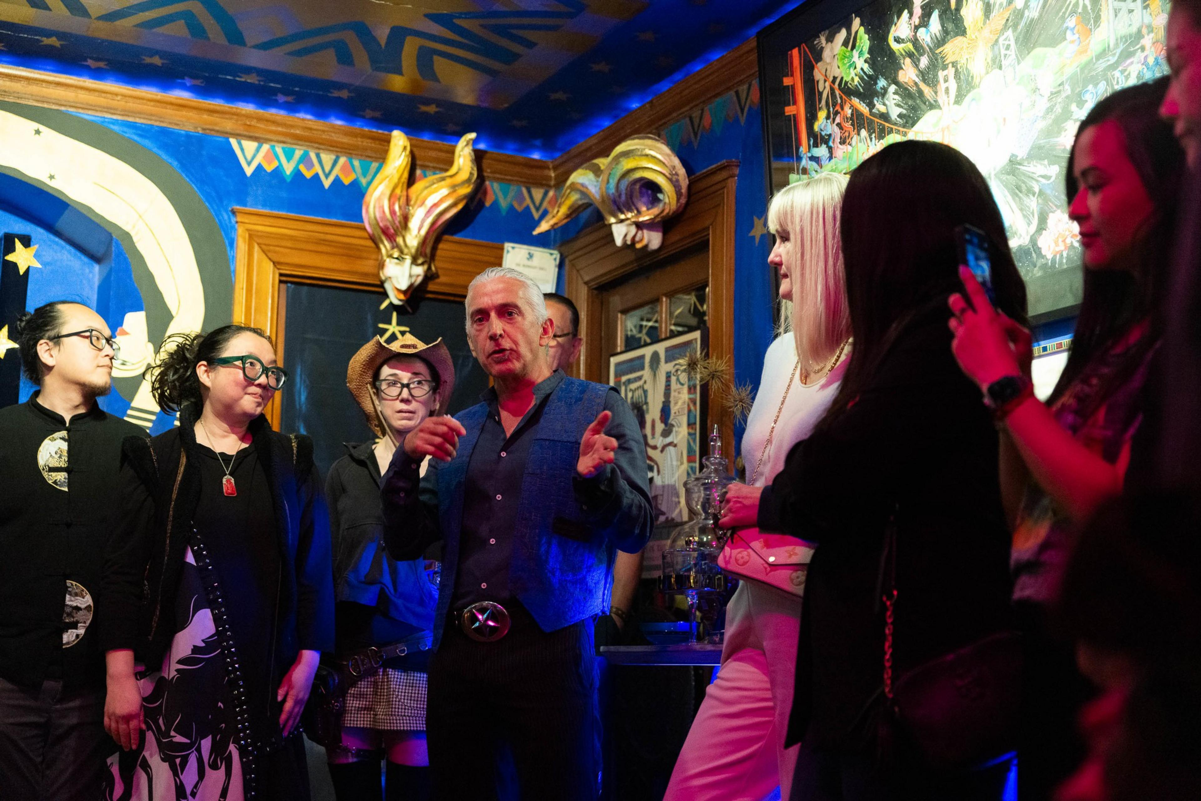 A group of people in a lively room with vibrant, carnival-like decor is focused on a man in the center speaking and gesturing.