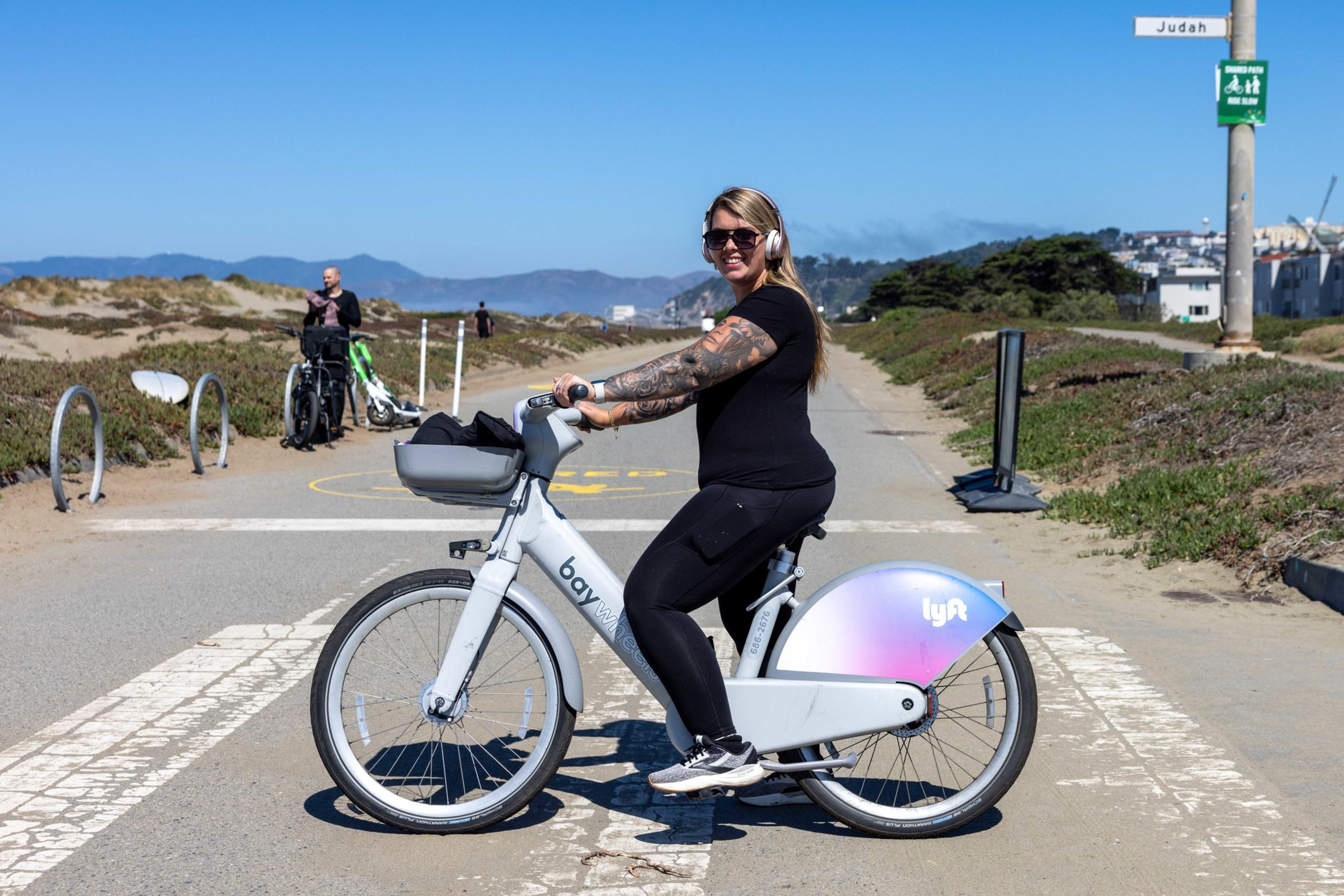 A woman wearing sunglasses and headphones rides a white Lyft bike on a sunny paved path near sandy dunes, with mountains in the background.