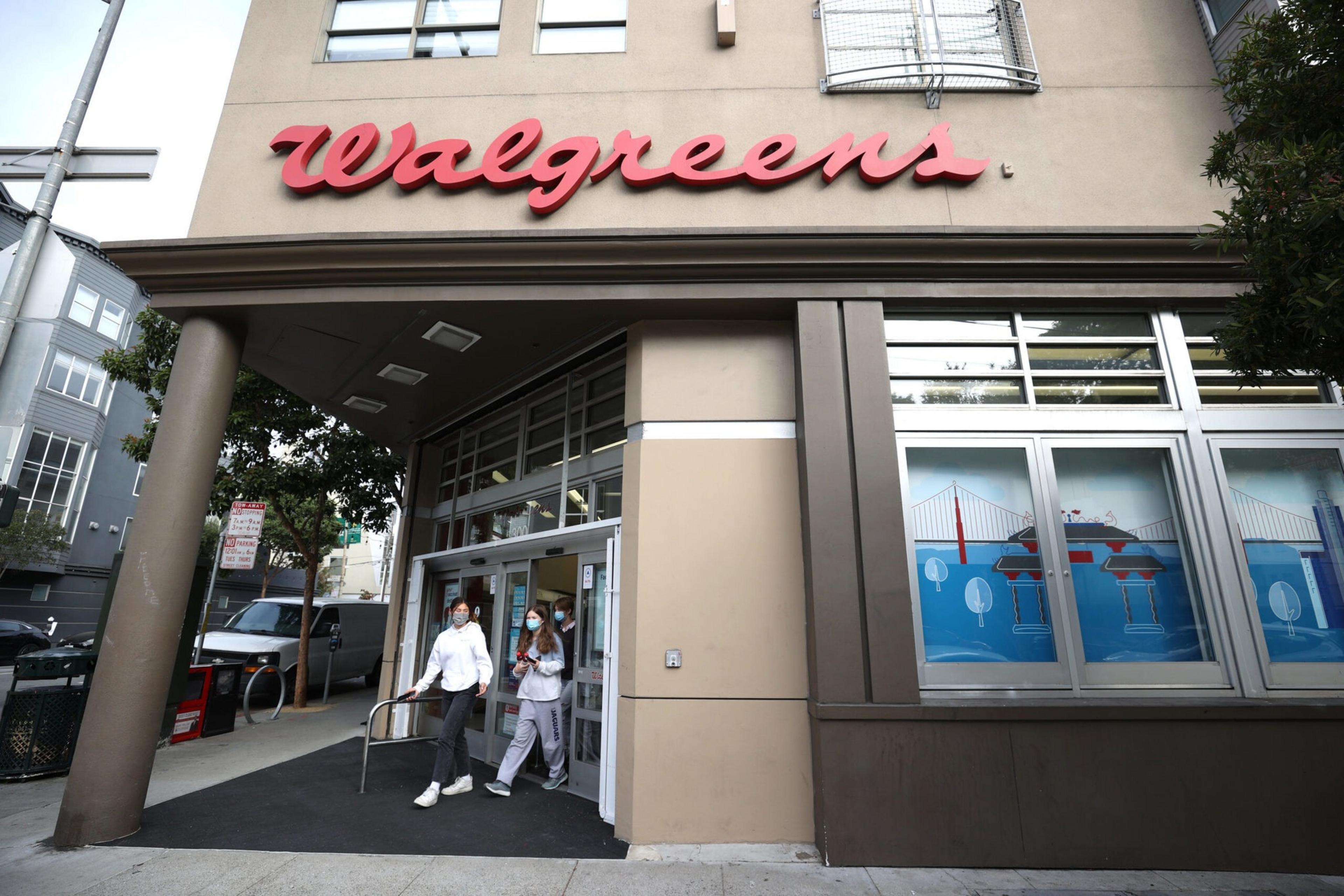 A Walgreens store entrance with two people wearing masks walking out, and windows displaying artwork of a red bridge over blue water.