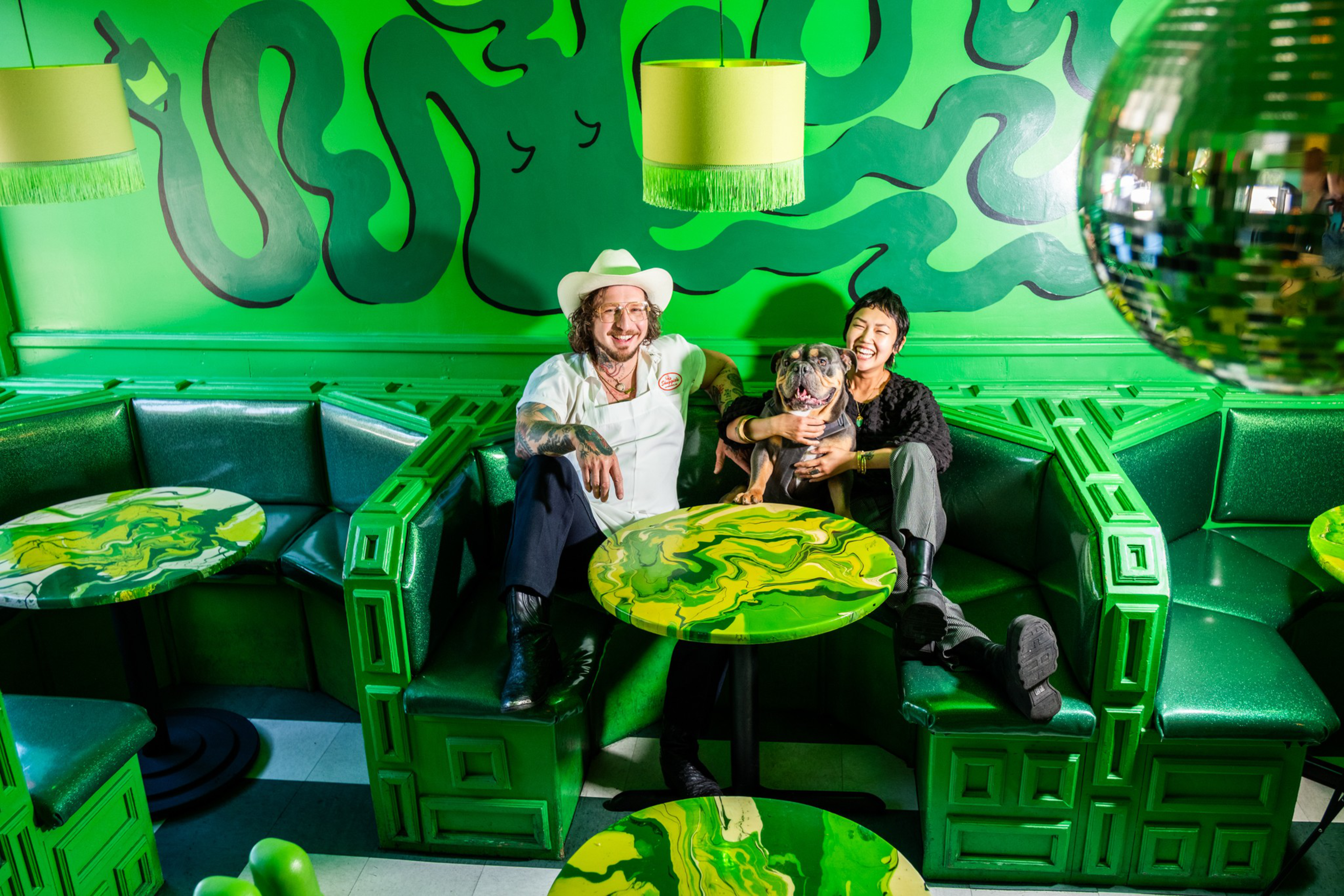 A man in a white hat and a woman smile while sitting on a green booth with a dog, surrounded by green tables and a swirling green wall mural.