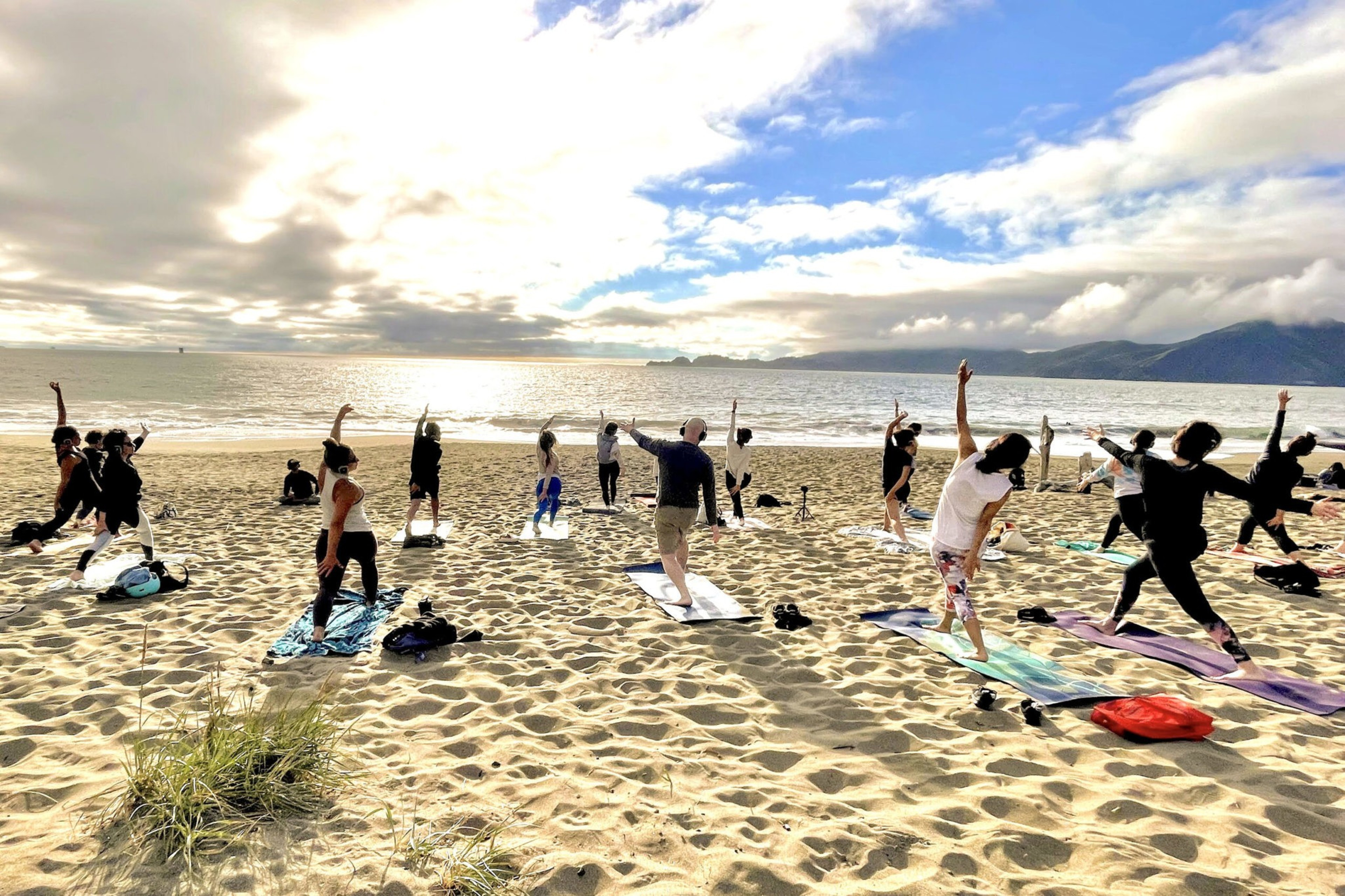 A group practicing yoga on a sunny beach with mountains in the distance.