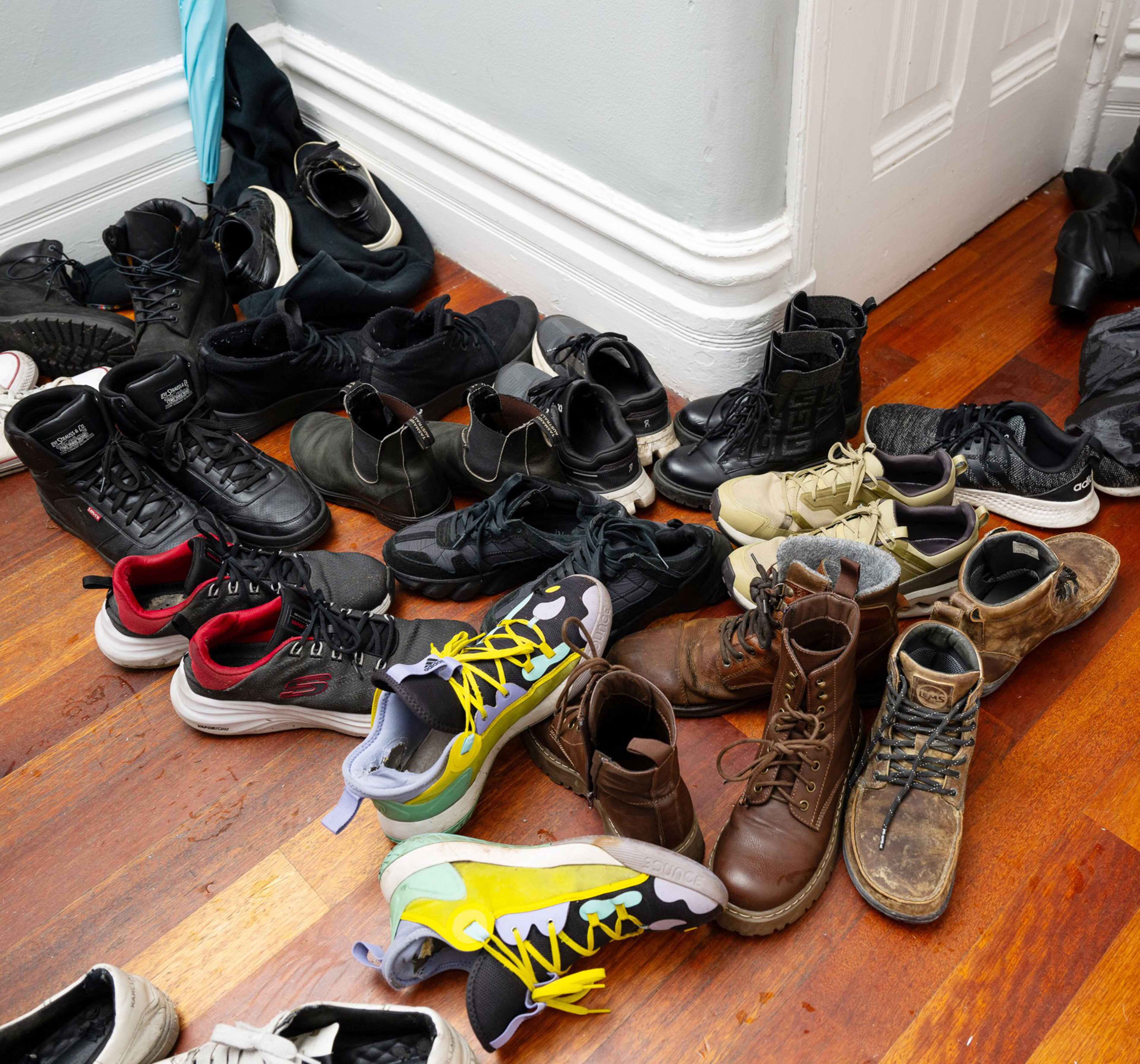 The image shows a cluttered collection of various shoes, including sneakers, boots, and casual footwear, scattered on a wooden floor near a white wall.