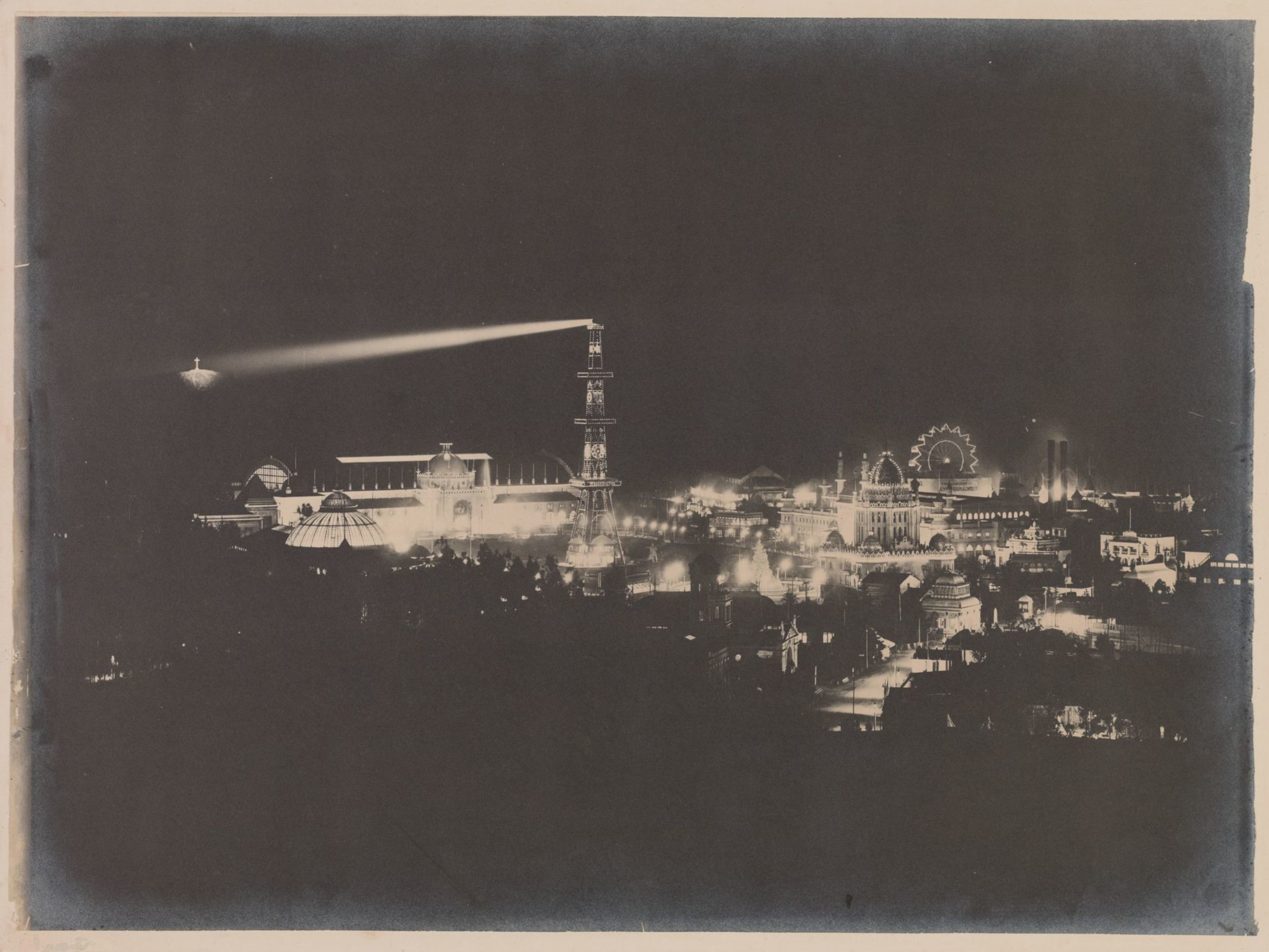 Brightly lit buildings, a tall illuminated tower, and a Ferris wheel can be seen at night, with a strong spotlight beaming from a floating platform overhead.