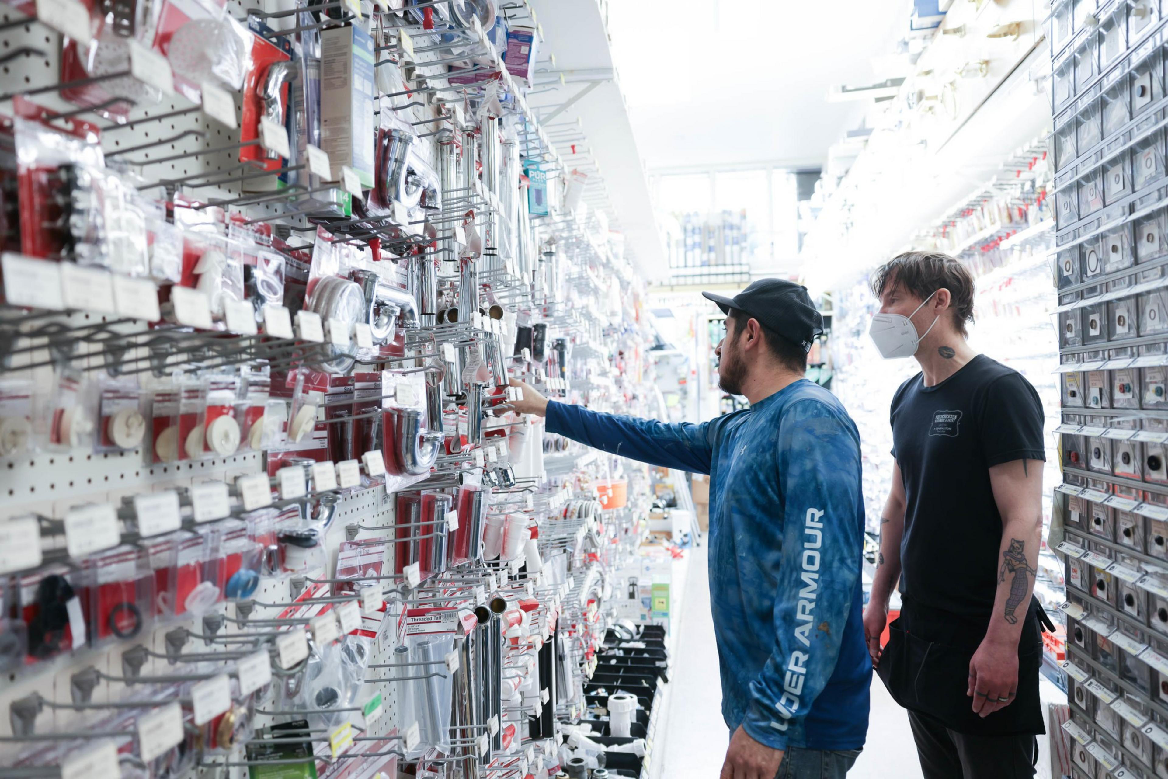 A man in a long sleeve blue shirt, glasses and caps reaches for item in a hardware store aisle as another man stands behind him