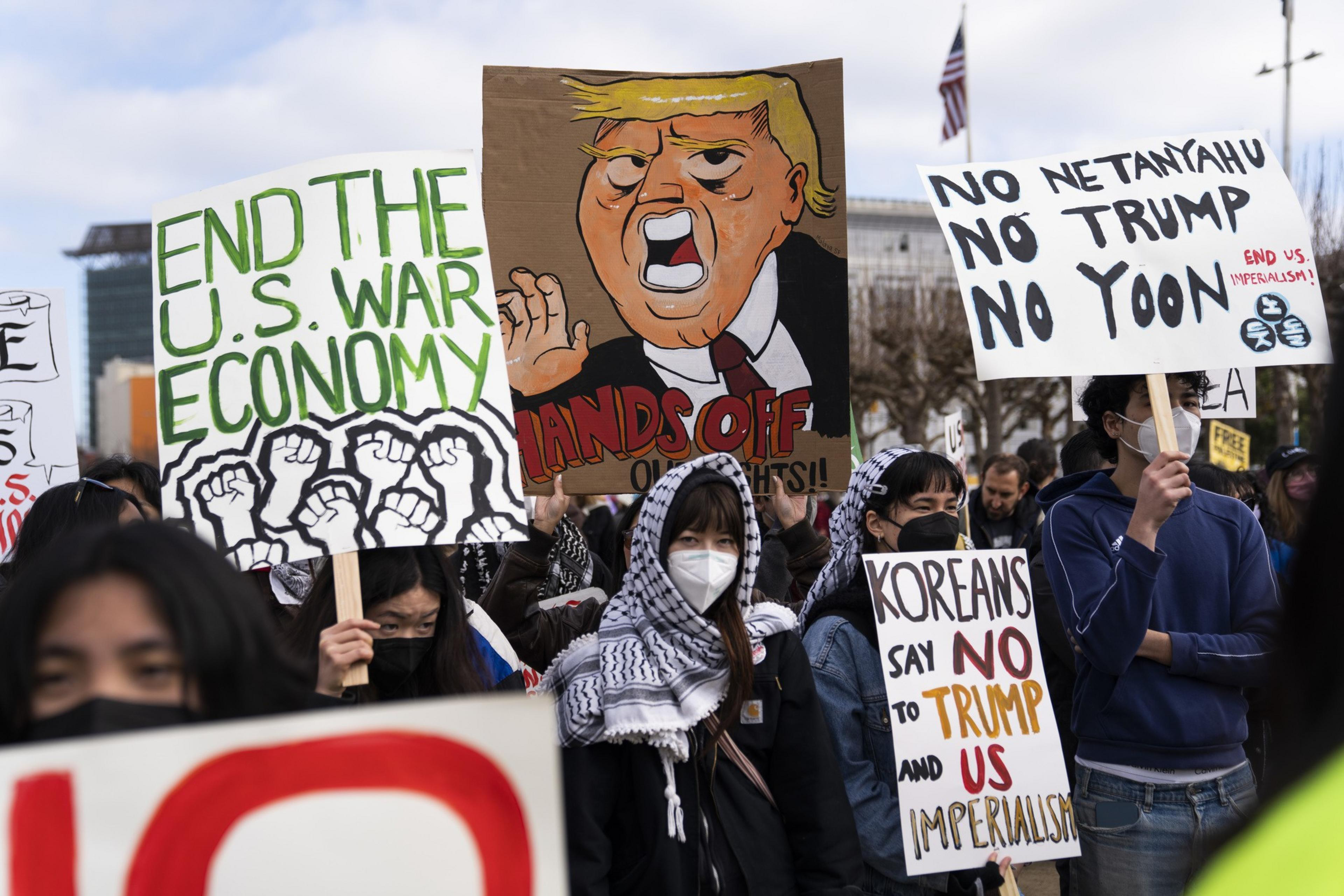 A group of protesters holds signs with messages opposing war, Trump, and imperialism. One sign features a caricature, while others emphasize ending the U.S. war economy.