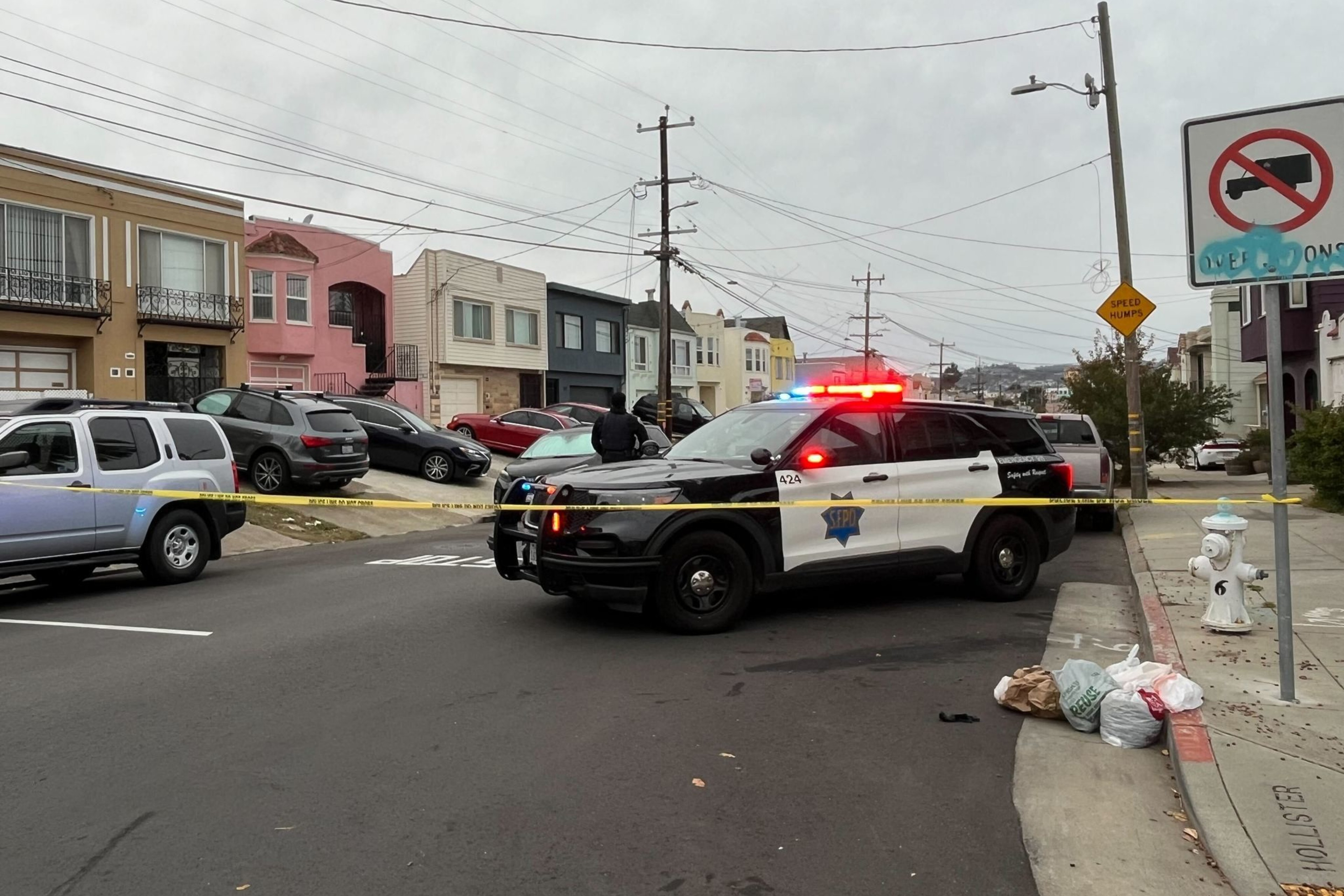 A police SUV with flashing lights blocks a street, alongside yellow caution tape. Nearby are parked cars, residential houses, and a few bags on the sidewalk.