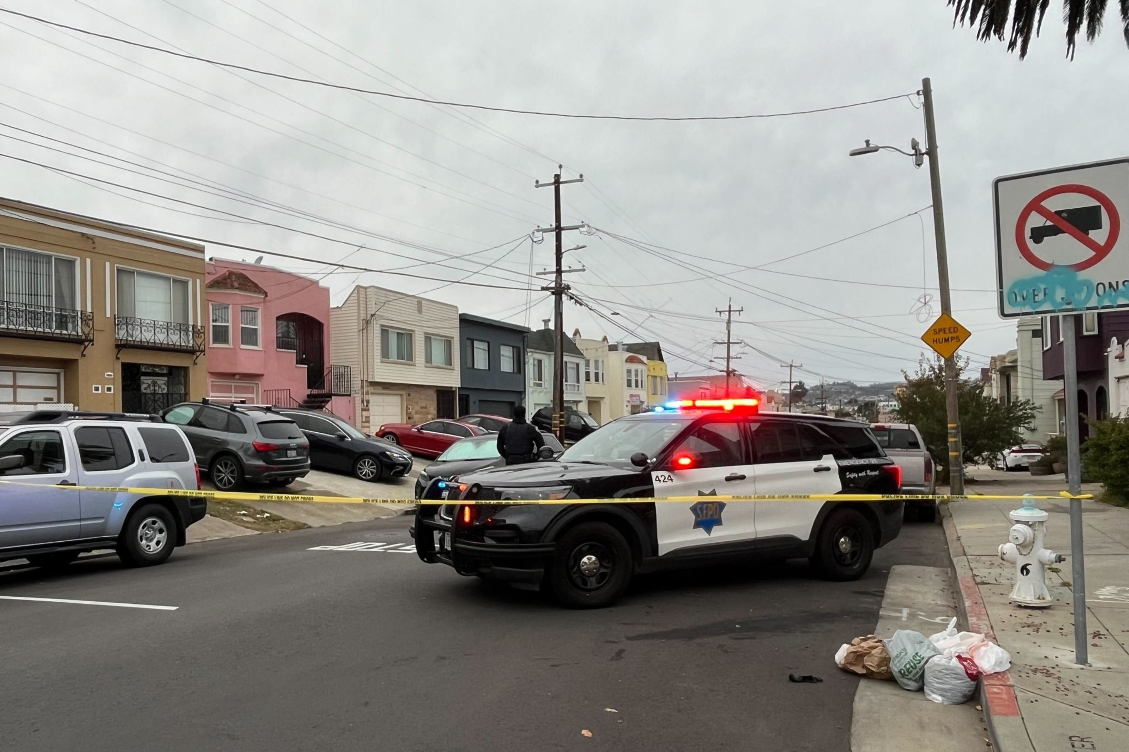A police SUV with flashing lights blocks a street, alongside yellow caution tape. Nearby are parked cars, residential houses, and a few bags on the sidewalk.
