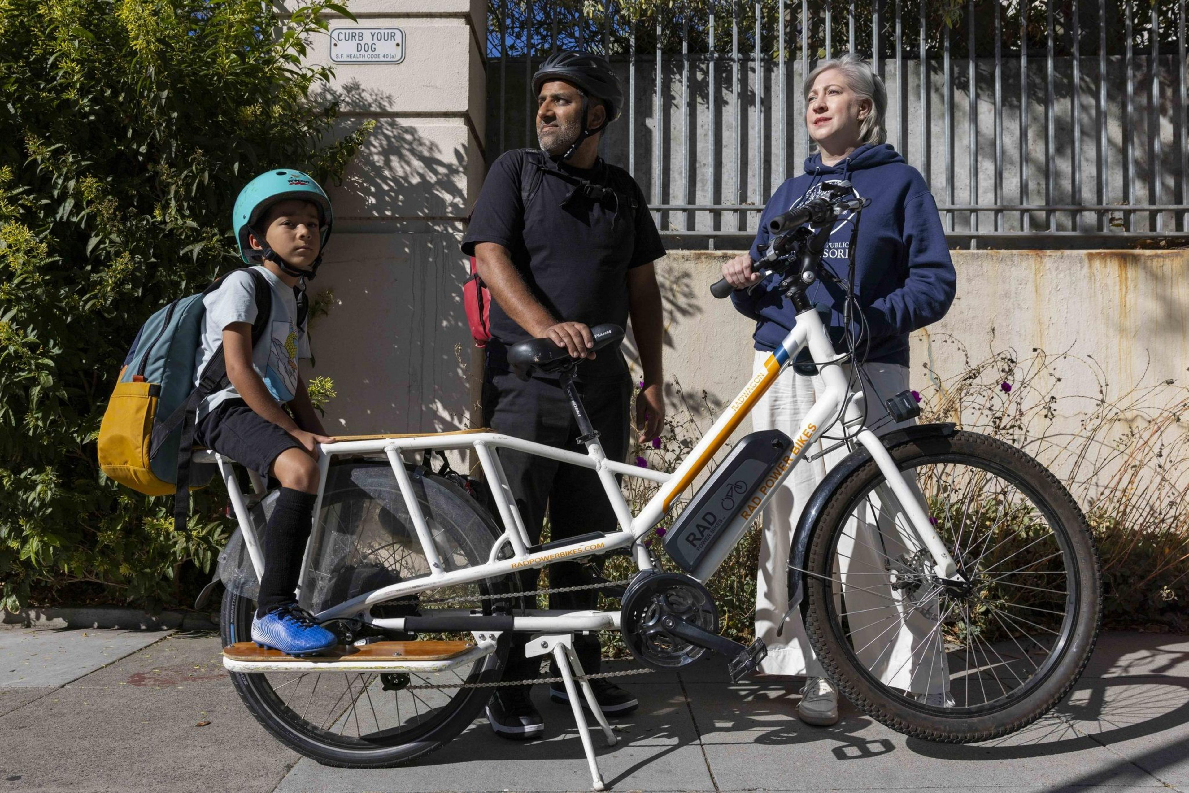A child in a blue helmet sits on a cargo bike, while two adults stand beside it, both wearing helmets. They appear to be on a sidewalk near a "Curb Your Dog" sign.