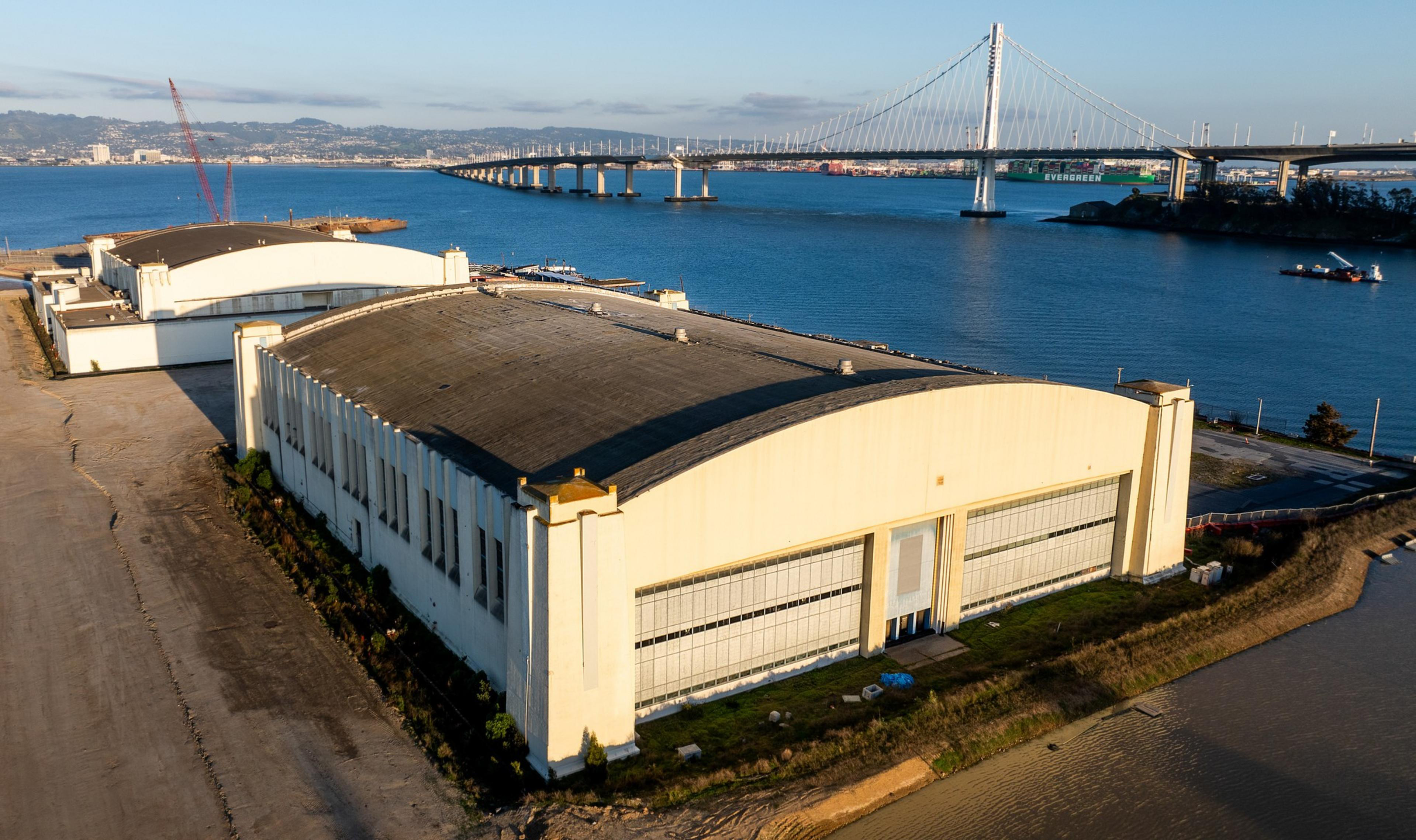 The image shows large warehouse-style buildings beside a body of water with a bridge in the background under a blue sky.