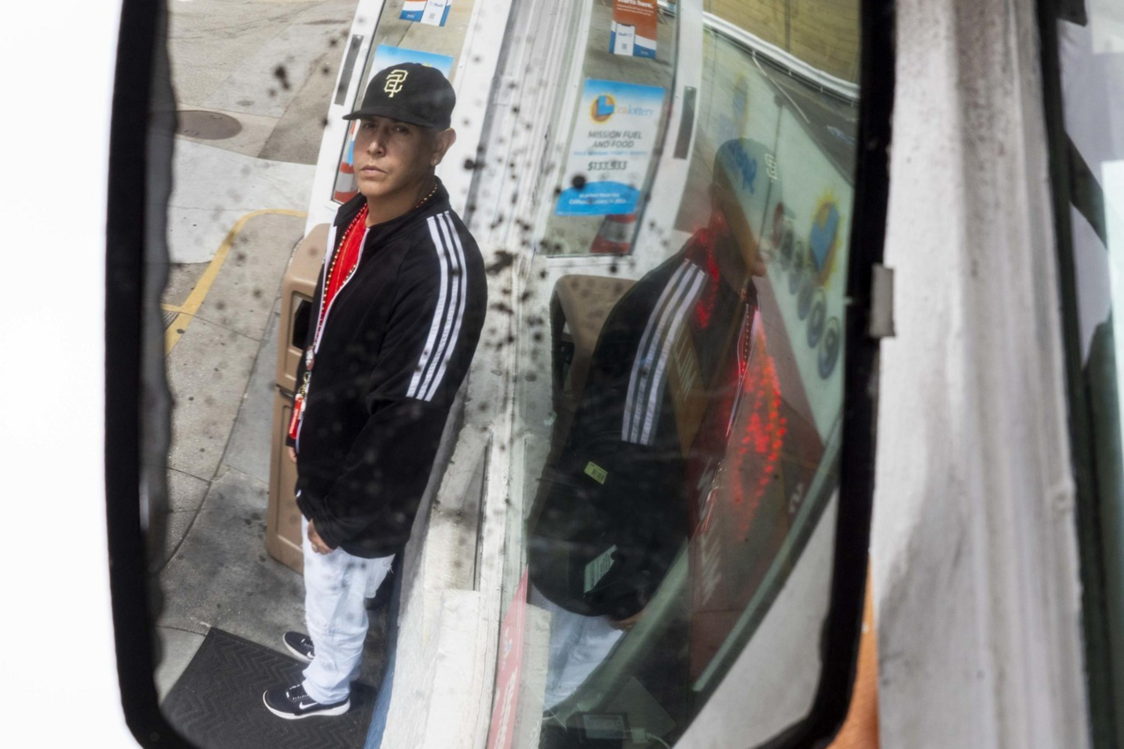A man is seen reflected in a dirty convex mirror, standing near a gas station window with signage about speedy Gulf Pay payments. He wears a black cap and jacket.