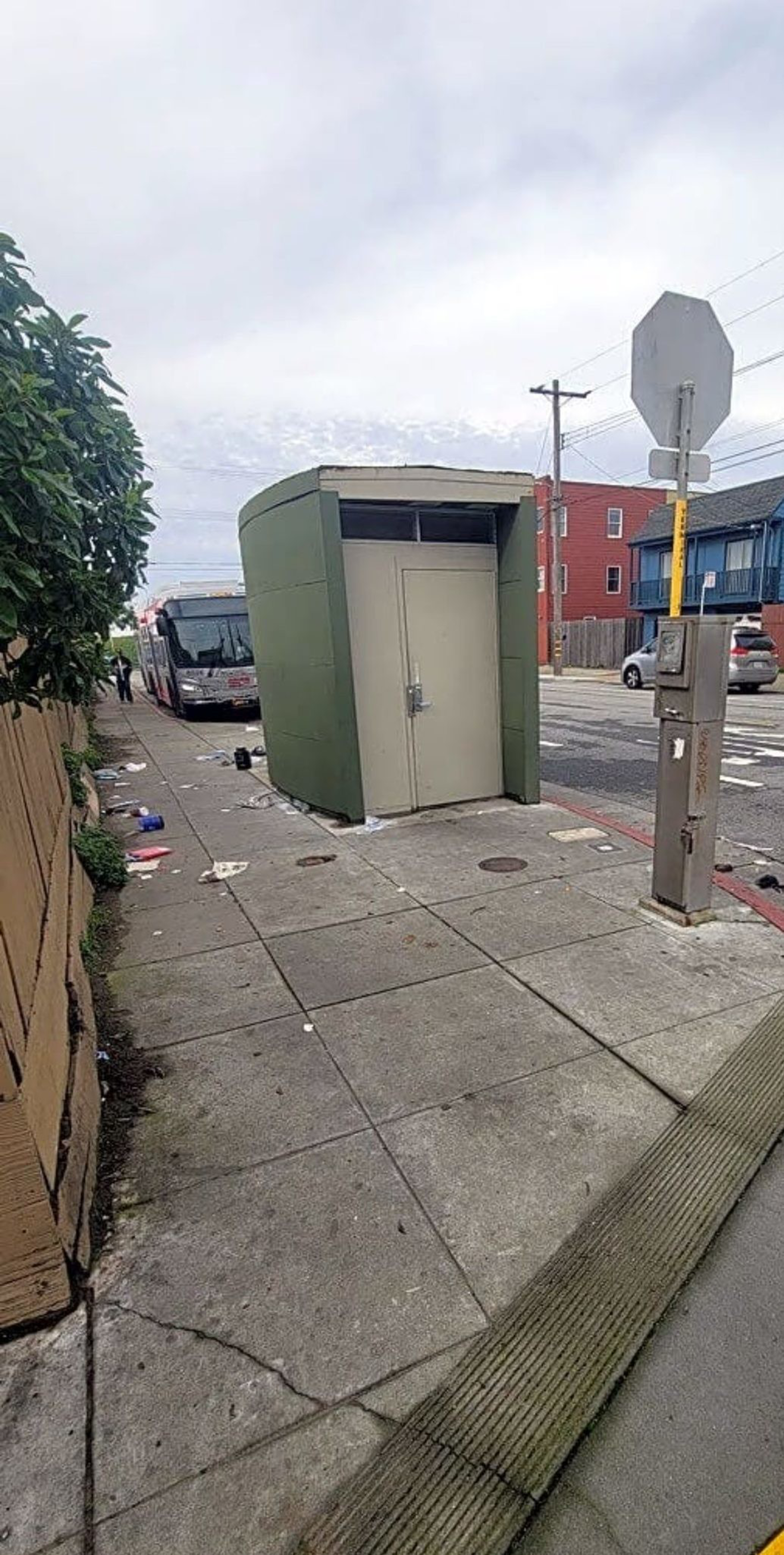 A green public restroom structure is on a littered sidewalk beside a road with a bus nearby. There's a utility box and some buildings in the background.