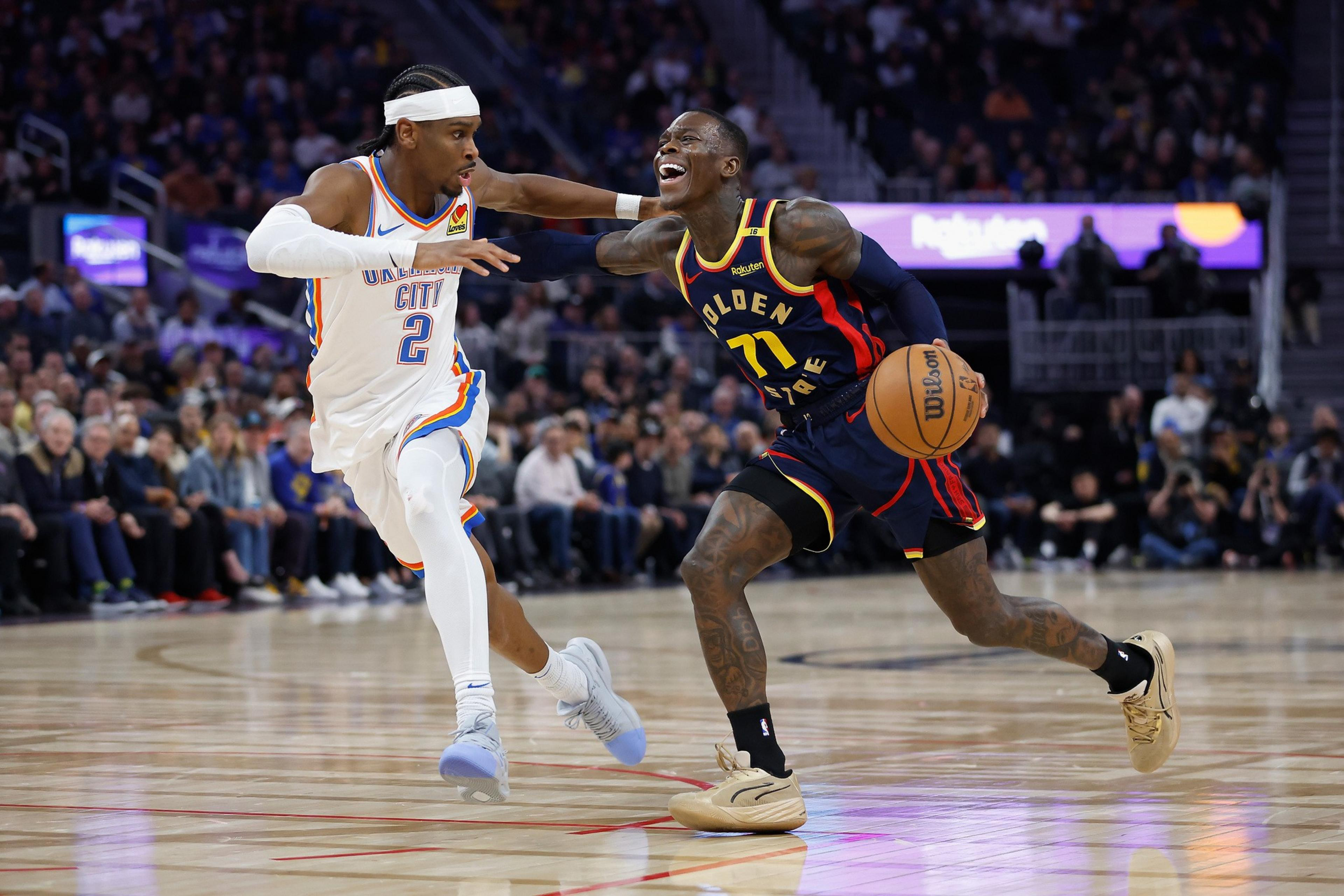 Two basketball players are in action on the court. One in a white Oklahoma City uniform guards the other in a dark Golden State uniform, who is dribbling the ball.