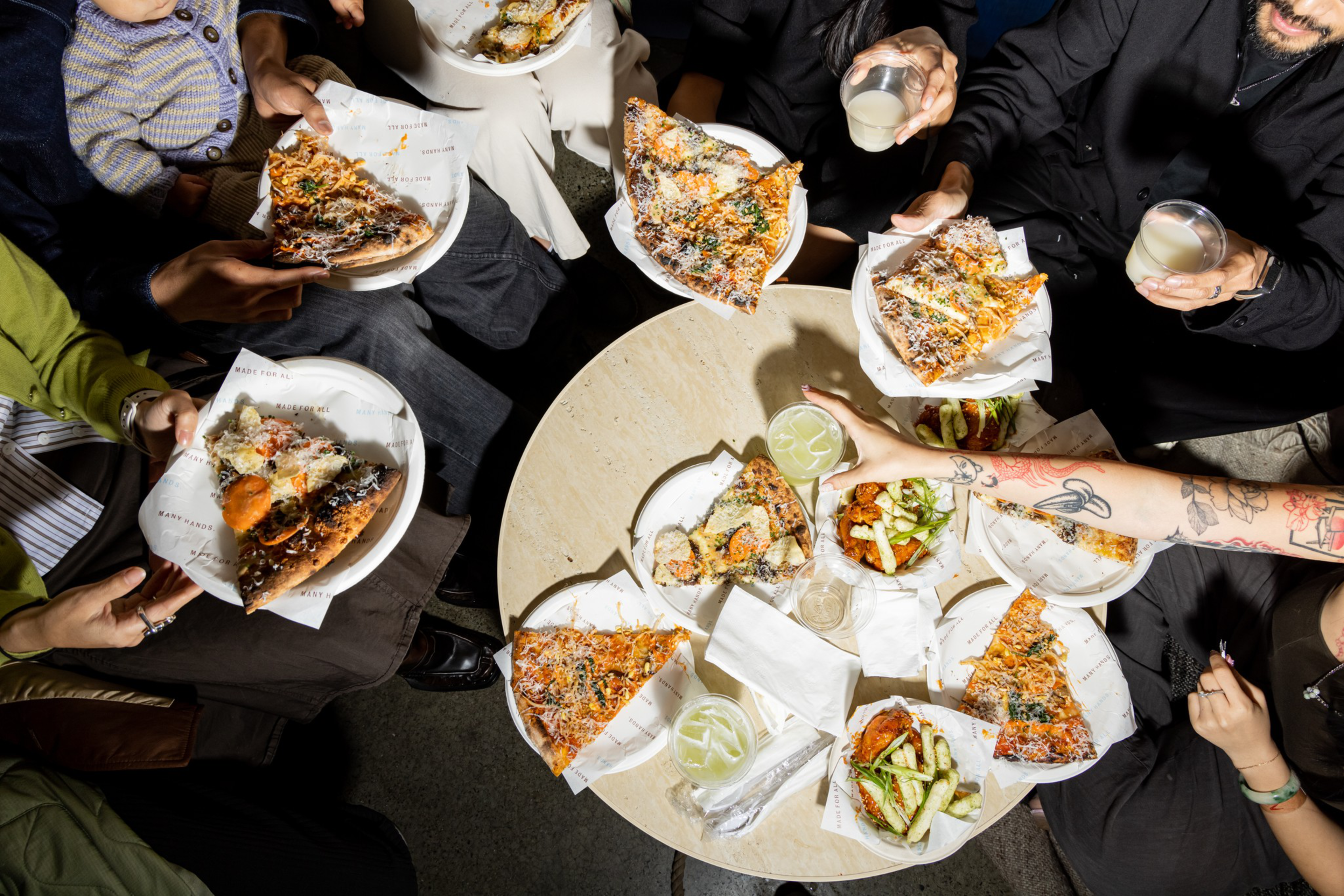 Six people are holding pizza slices and drinks around a small round table with more pizza slices, glasses, and plates of food.
