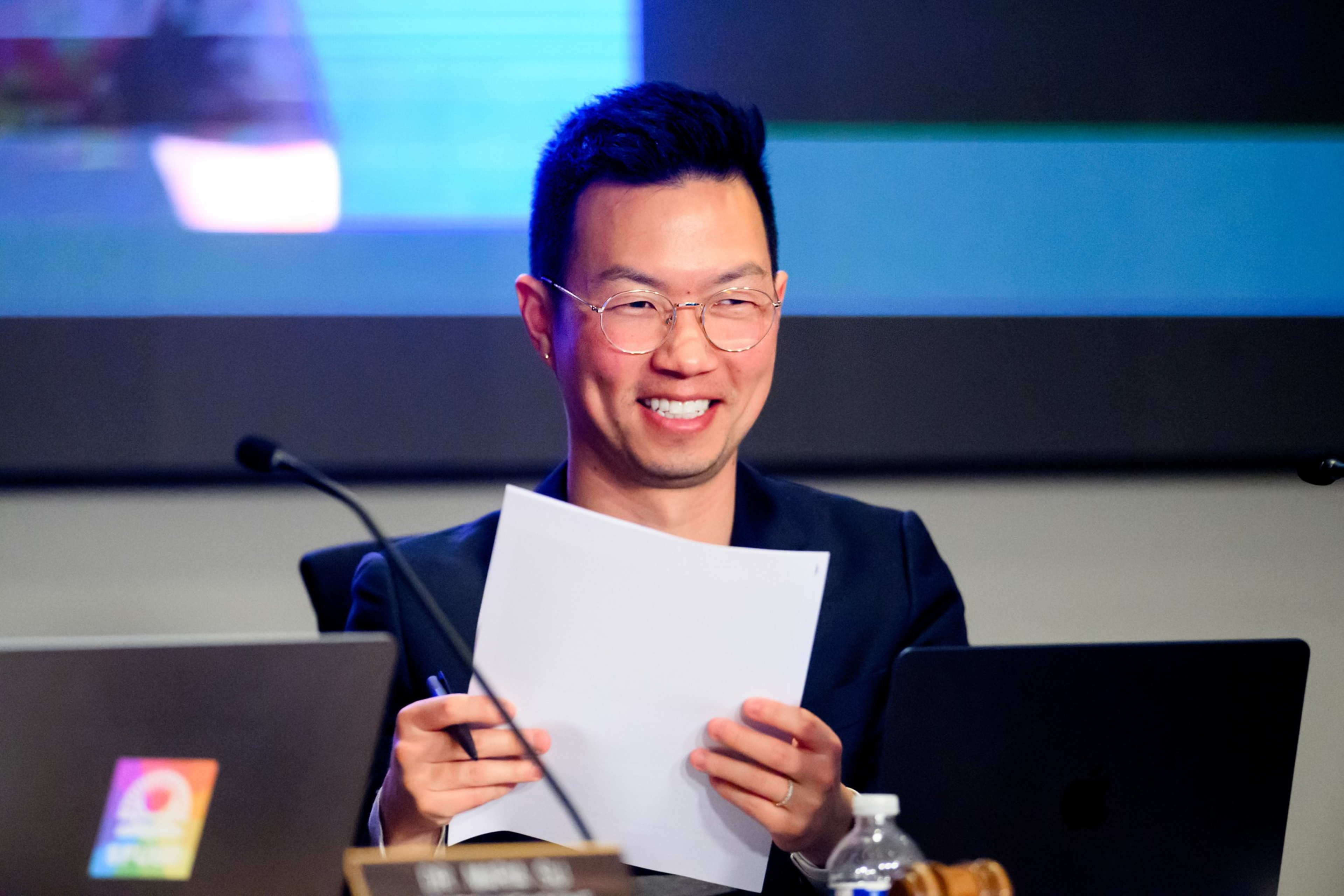 A man with glasses smiles while holding papers, sitting between two open laptops and a water bottle in a meeting or presentation setting.
