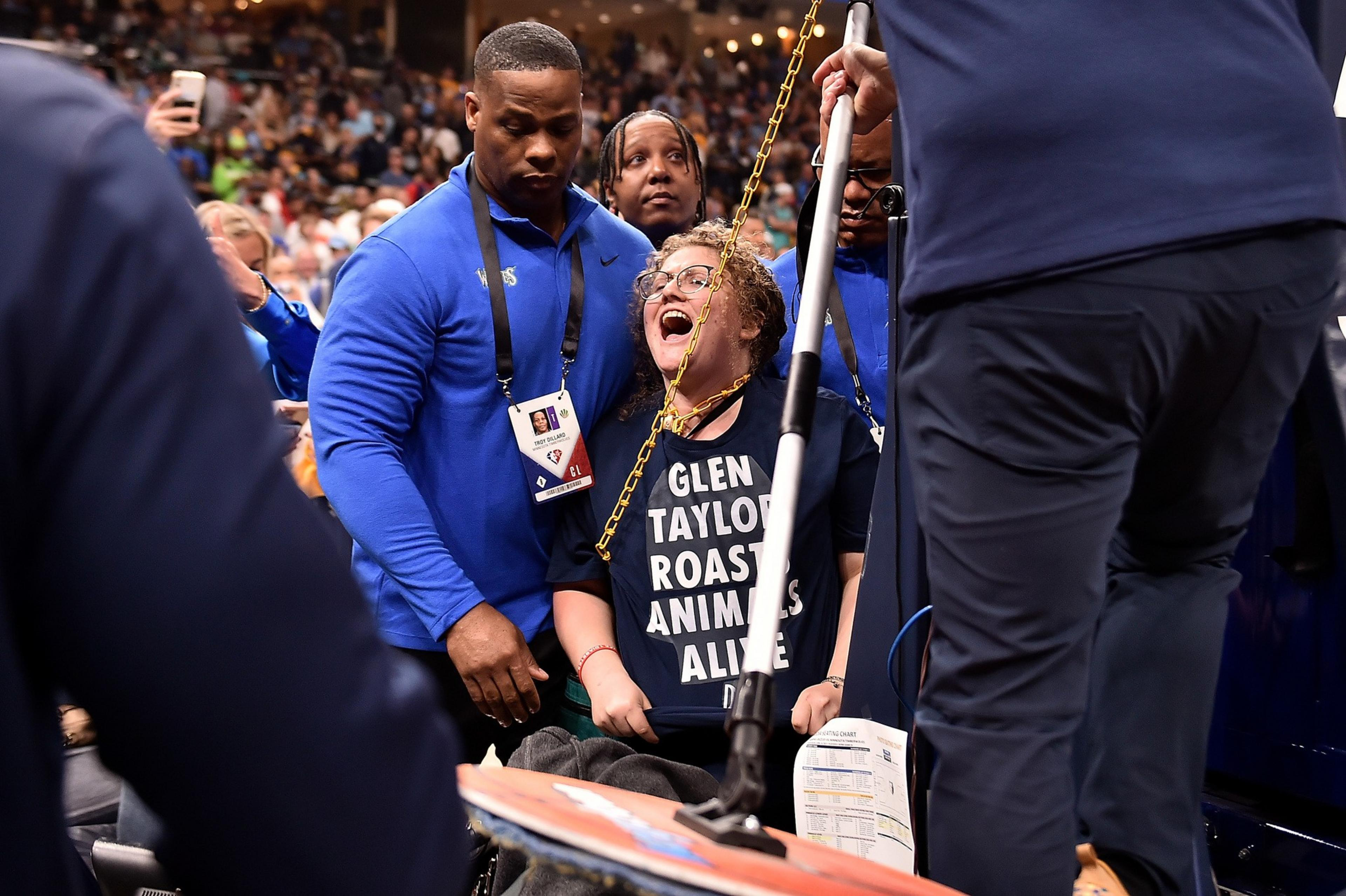 A person in a black shirt with white text is being restrained by security personnel. The scene appears to take place in a crowded venue.