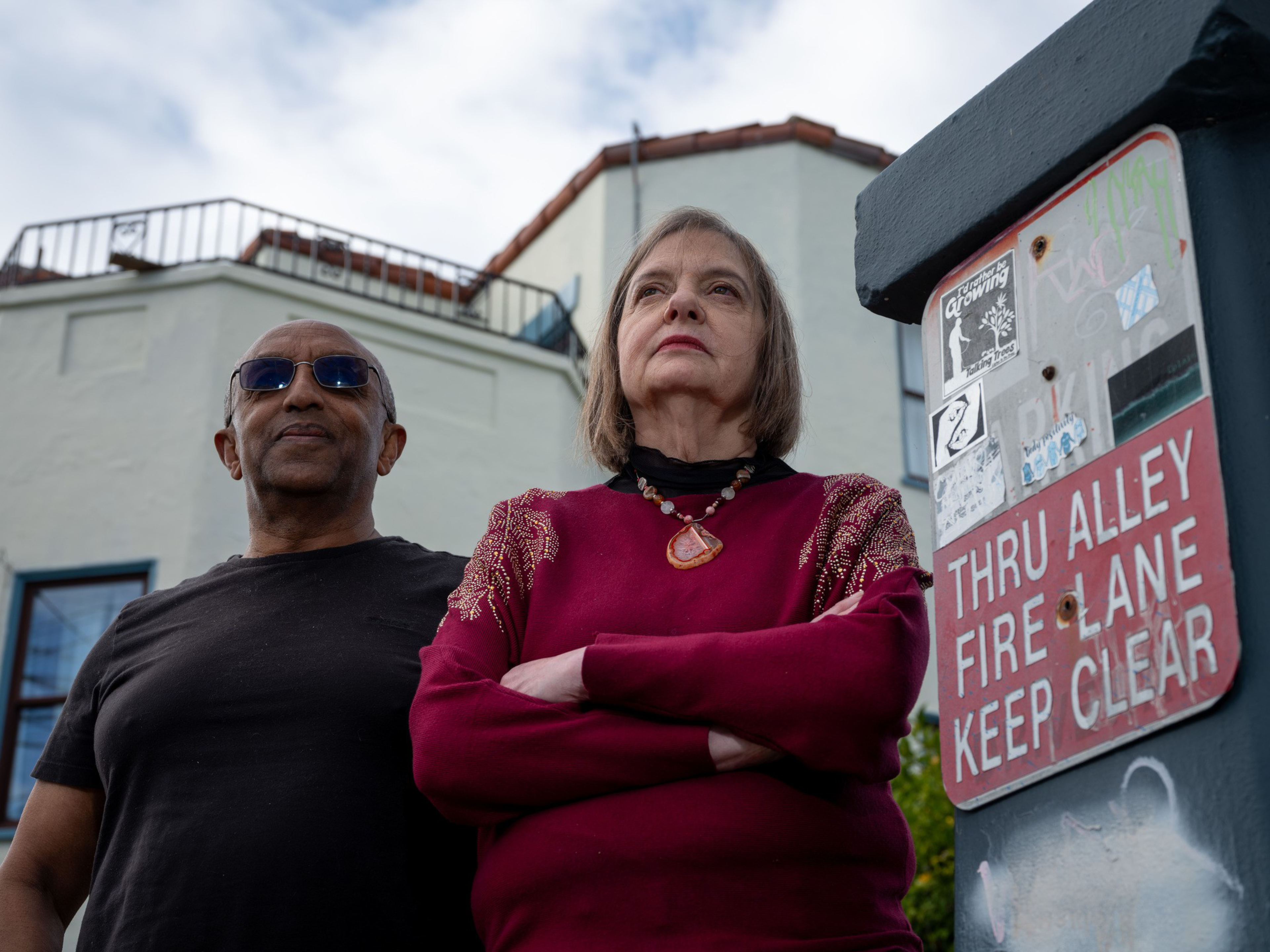 A serious-looking older woman in a red sweater and necklace stands with arms crossed next to a man in sunglasses and a dark shirt by a fire lane sign.
