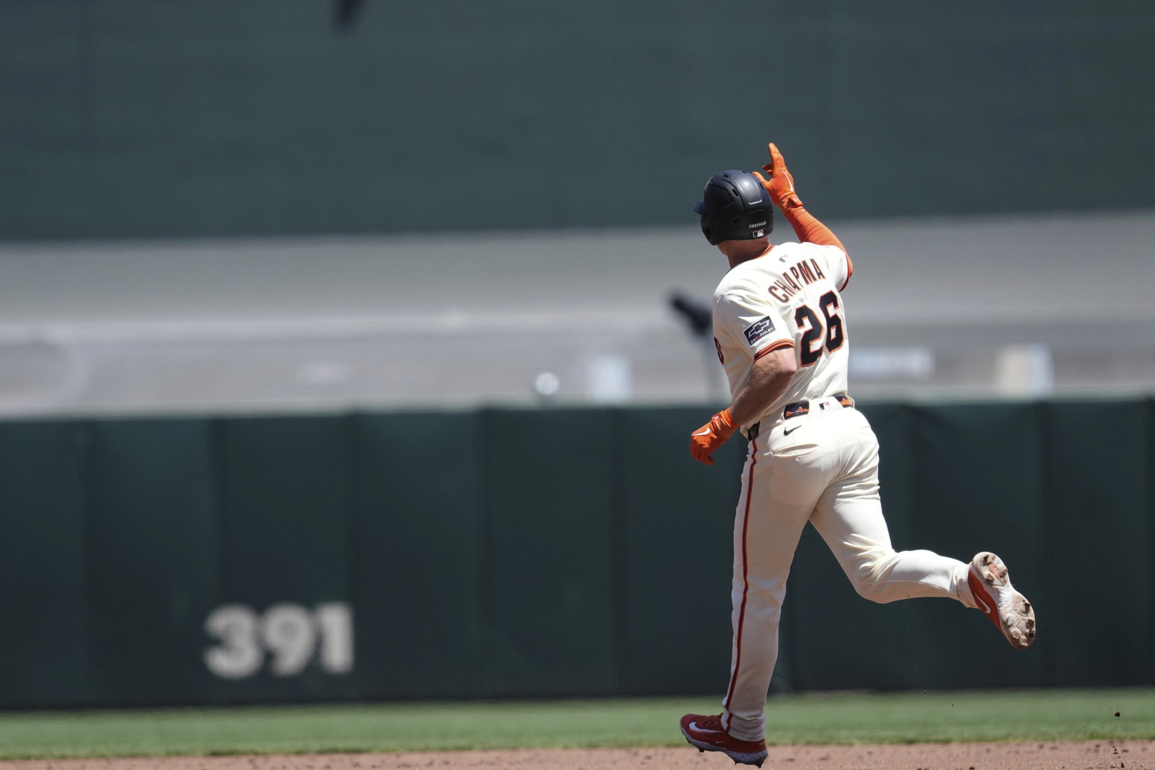 A baseball player in a white uniform runs on the field, pointing upwards with one hand. His jersey has the number 26, and the scoreboard reads &quot;391&quot; in the background.