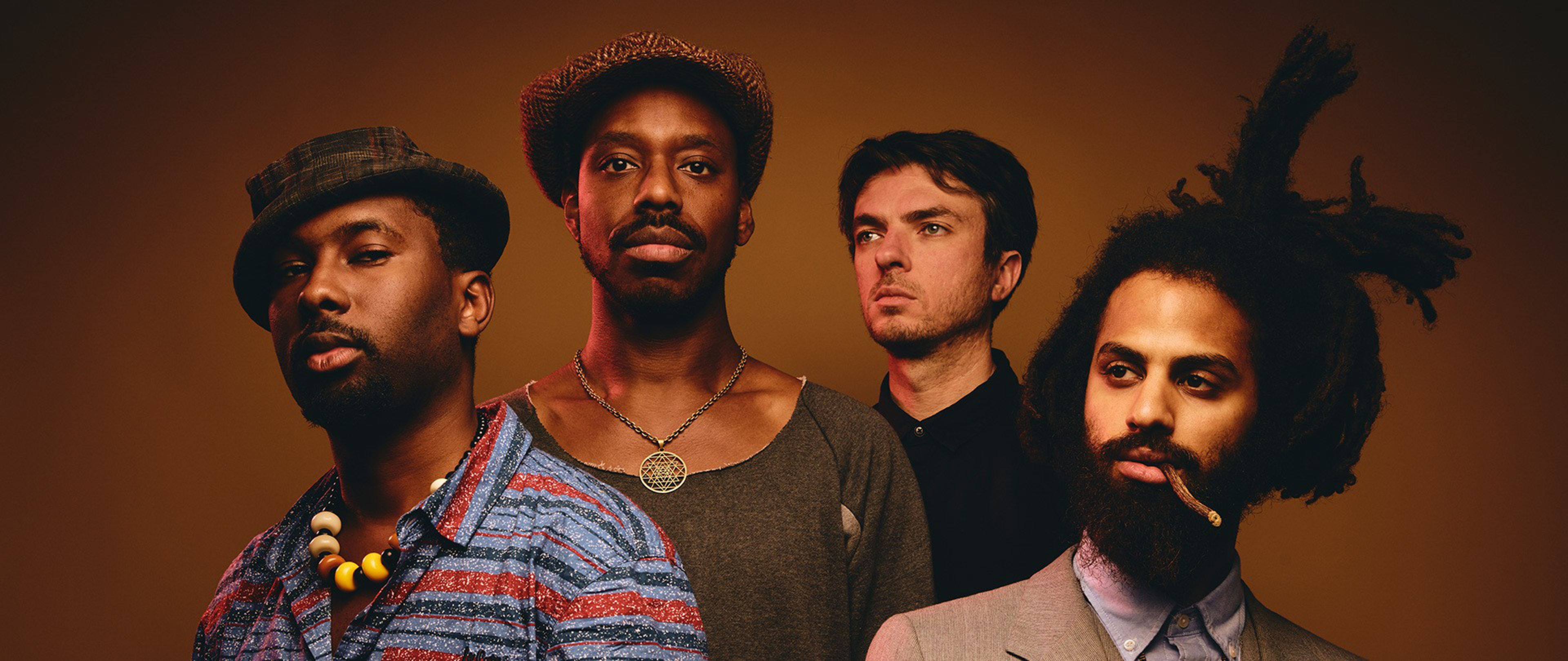 Four men stand against a dark background; two wear hats and casual clothes, one wears a black shirt, and one with dreadlocks smokes a cigarillo.