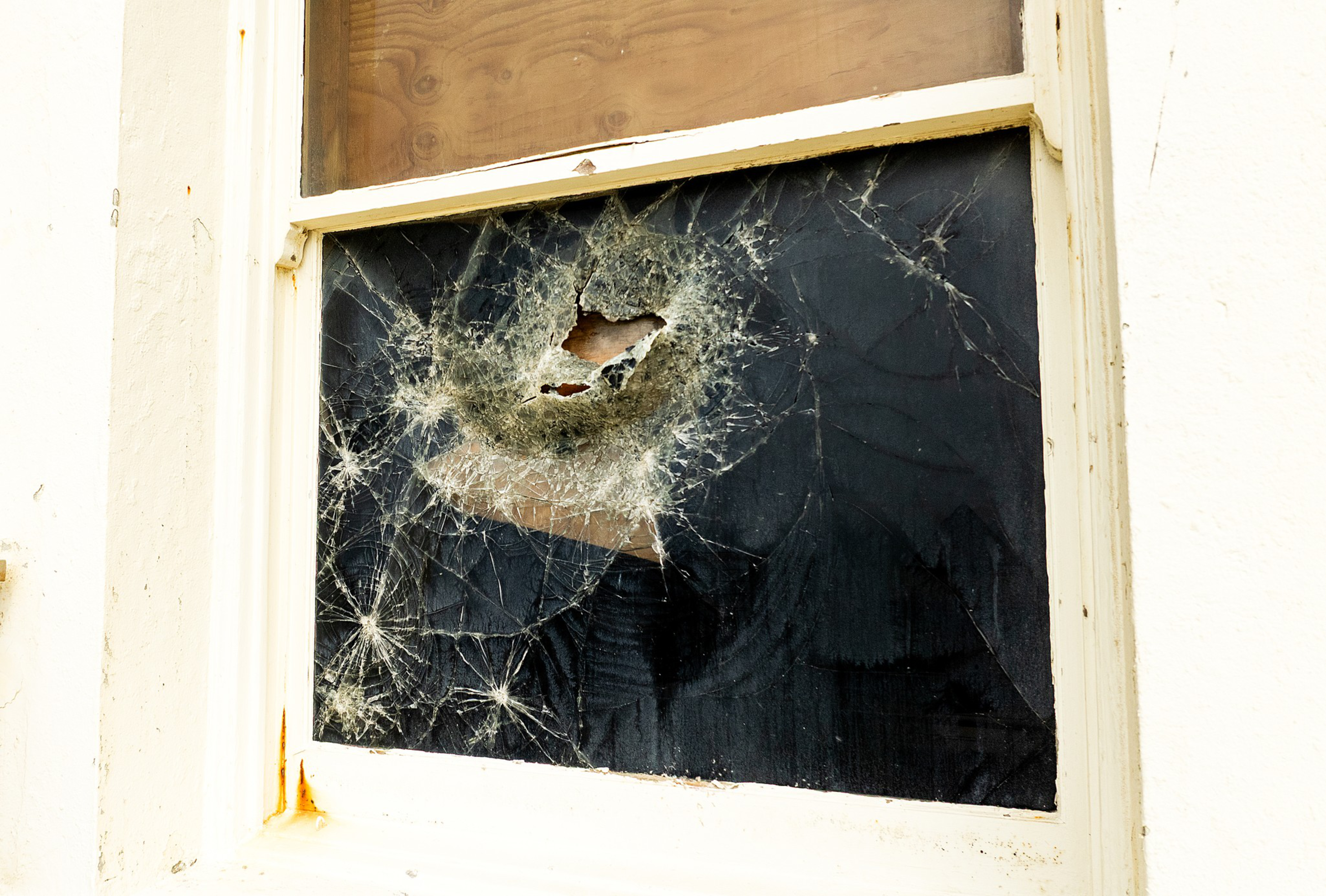 A window with a large, circular hole in the glass is surrounded by spiderweb-like cracks, all set in a white-painted frame.