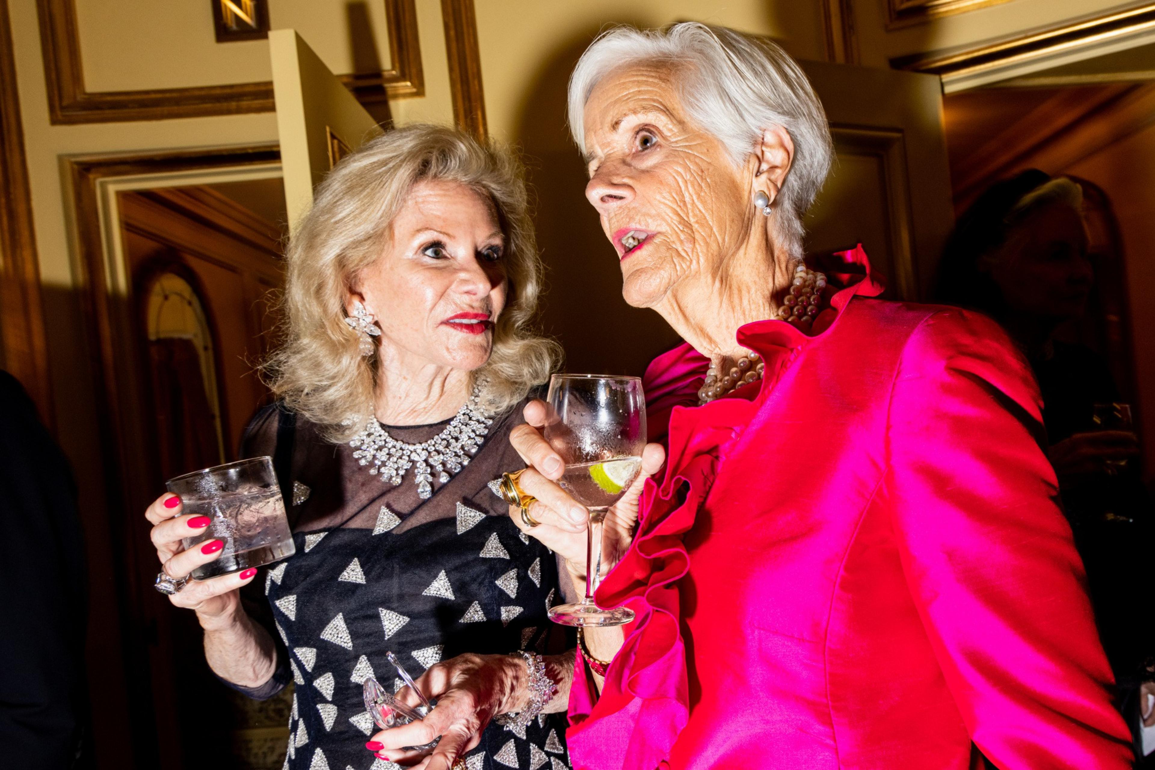 Two elderly women in elegant dresses hold drinks while engaging in an animated conversation at a formal indoor event.