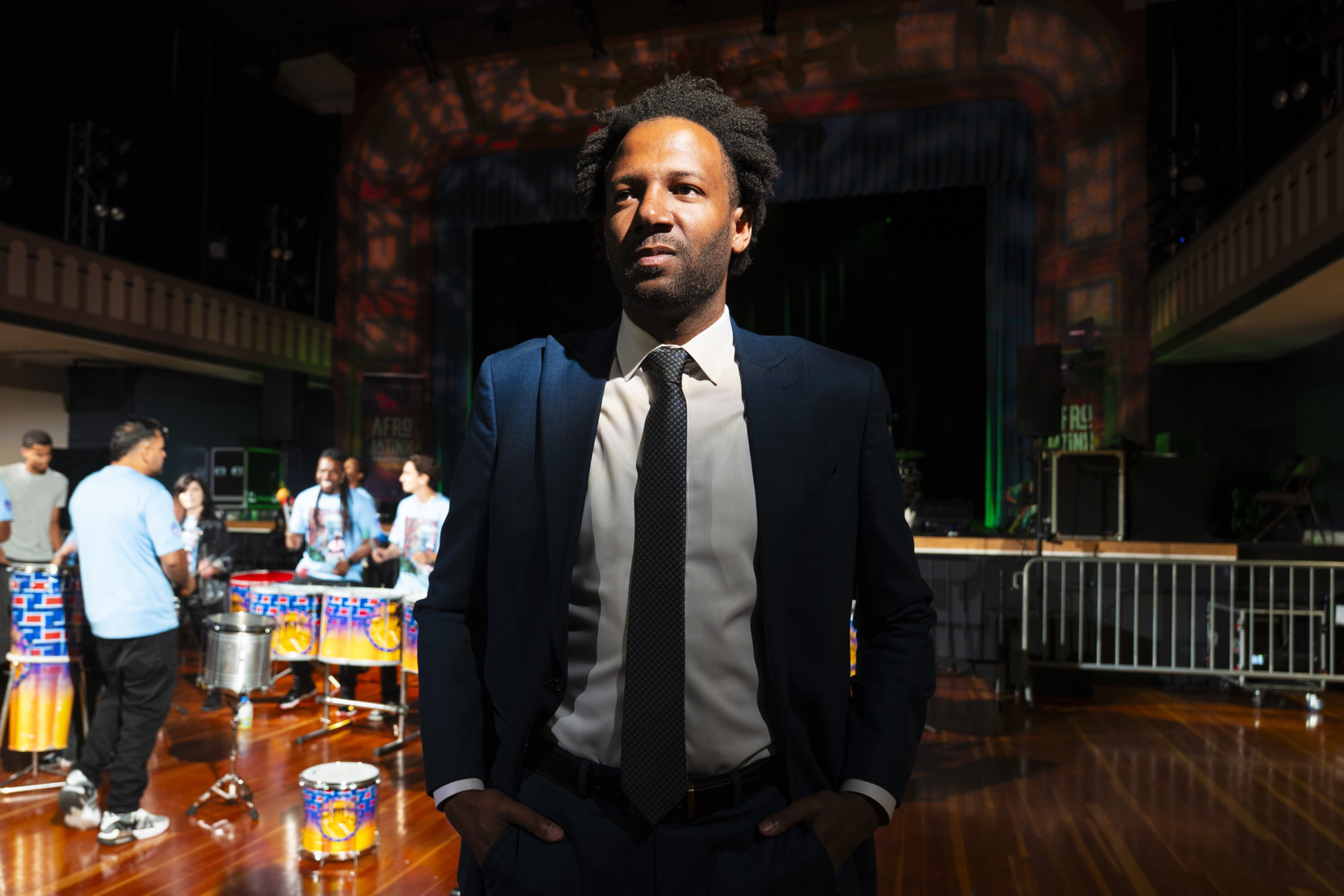 A man in a dark suit and tie stands confidently with hands in pockets in a brightly lit room where musicians play colorful drums in the background.