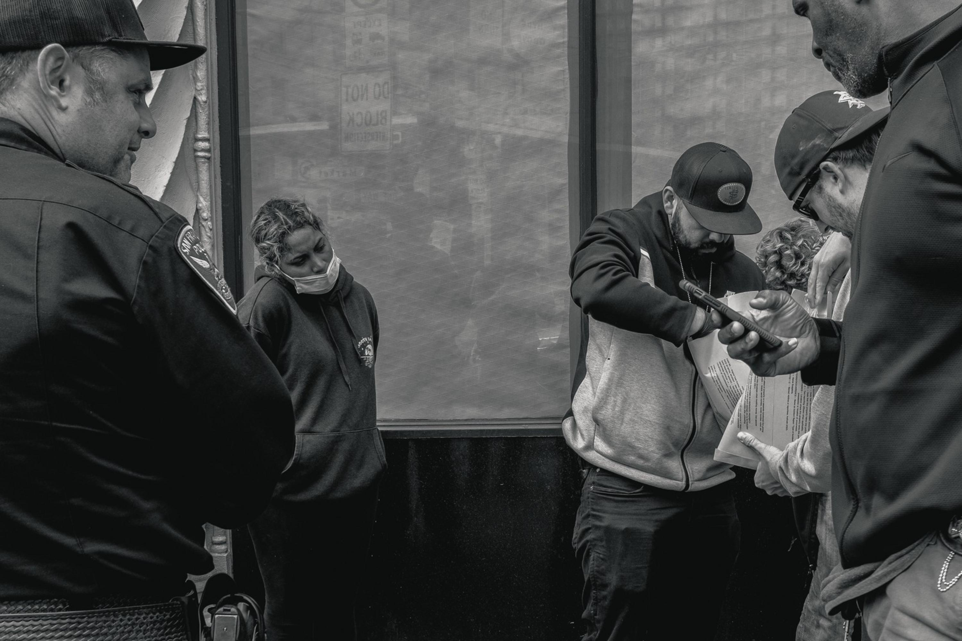 A group of people stand on a sidewalk, with one person appearing to show something on a phone to another. A masked woman leans against the wall nearby.