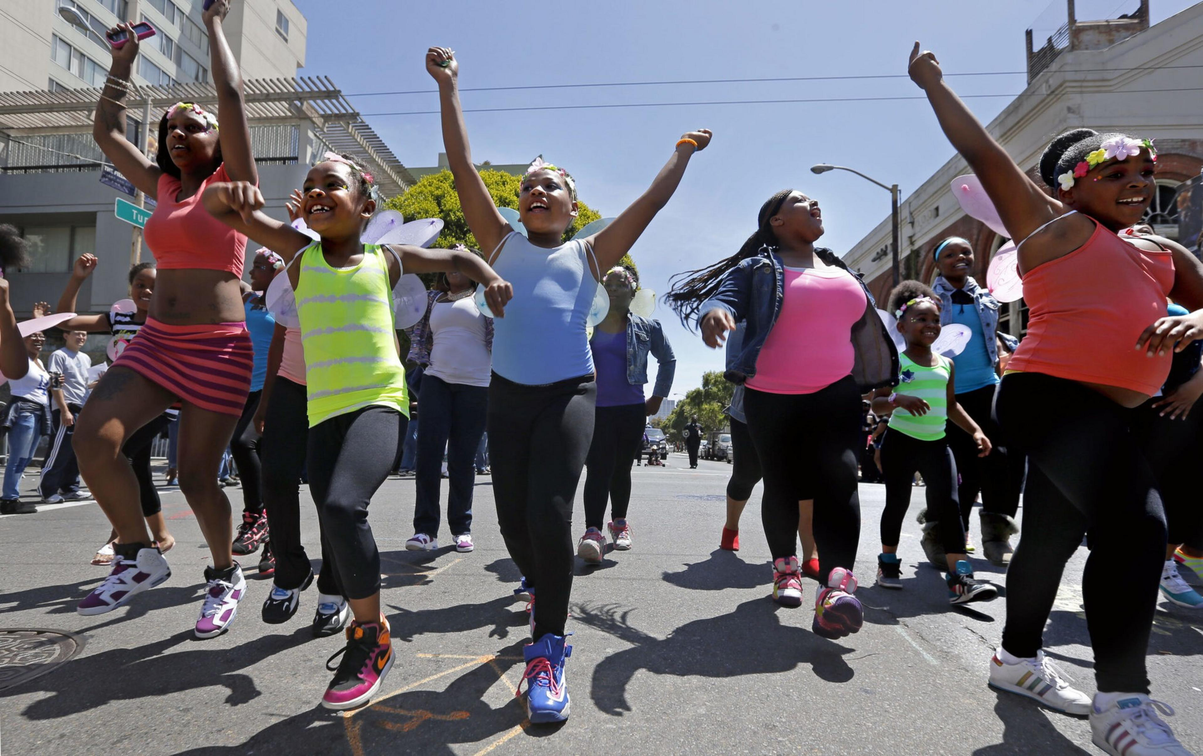 A group of girls wearing colorful clothes and fairy wings are joyfully dancing together on a sunny street, raising their arms in celebration.