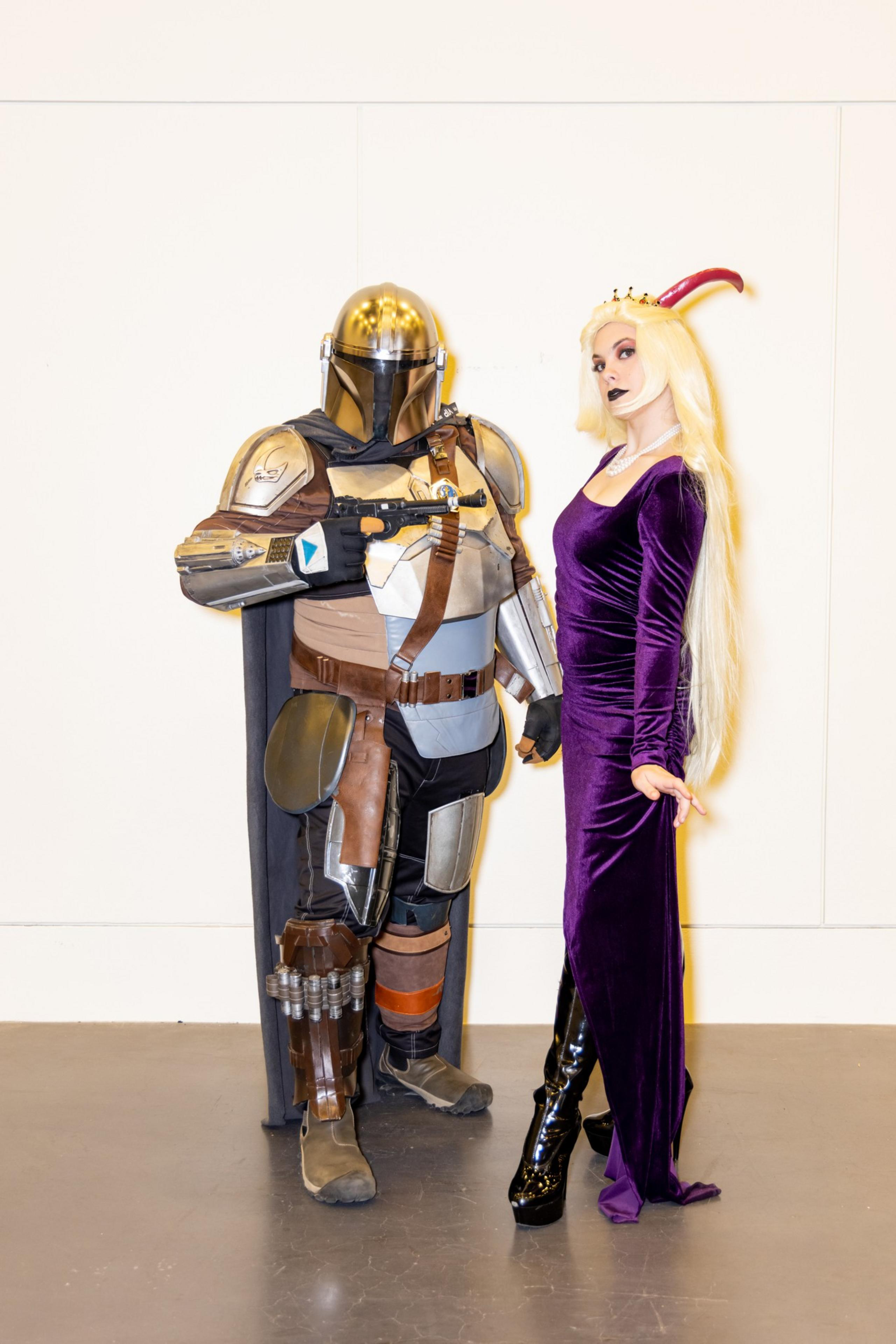 A person in detailed sci-fi armor stands next to another in a long purple dress and crown, both posing confidently against a plain background.