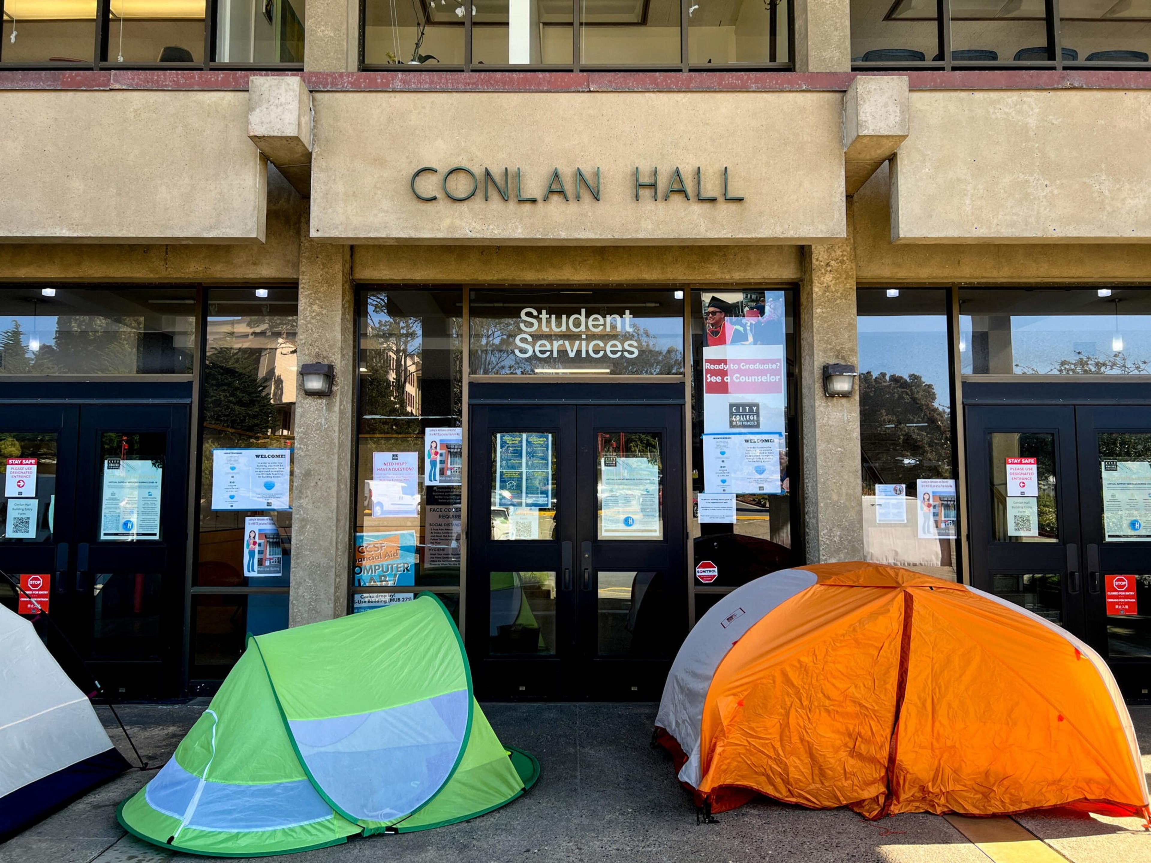 Three tents, green, orange, and gray, are set up outside the entrance of Conlan Hall, which houses Student Services, with various notices posted on the doors.