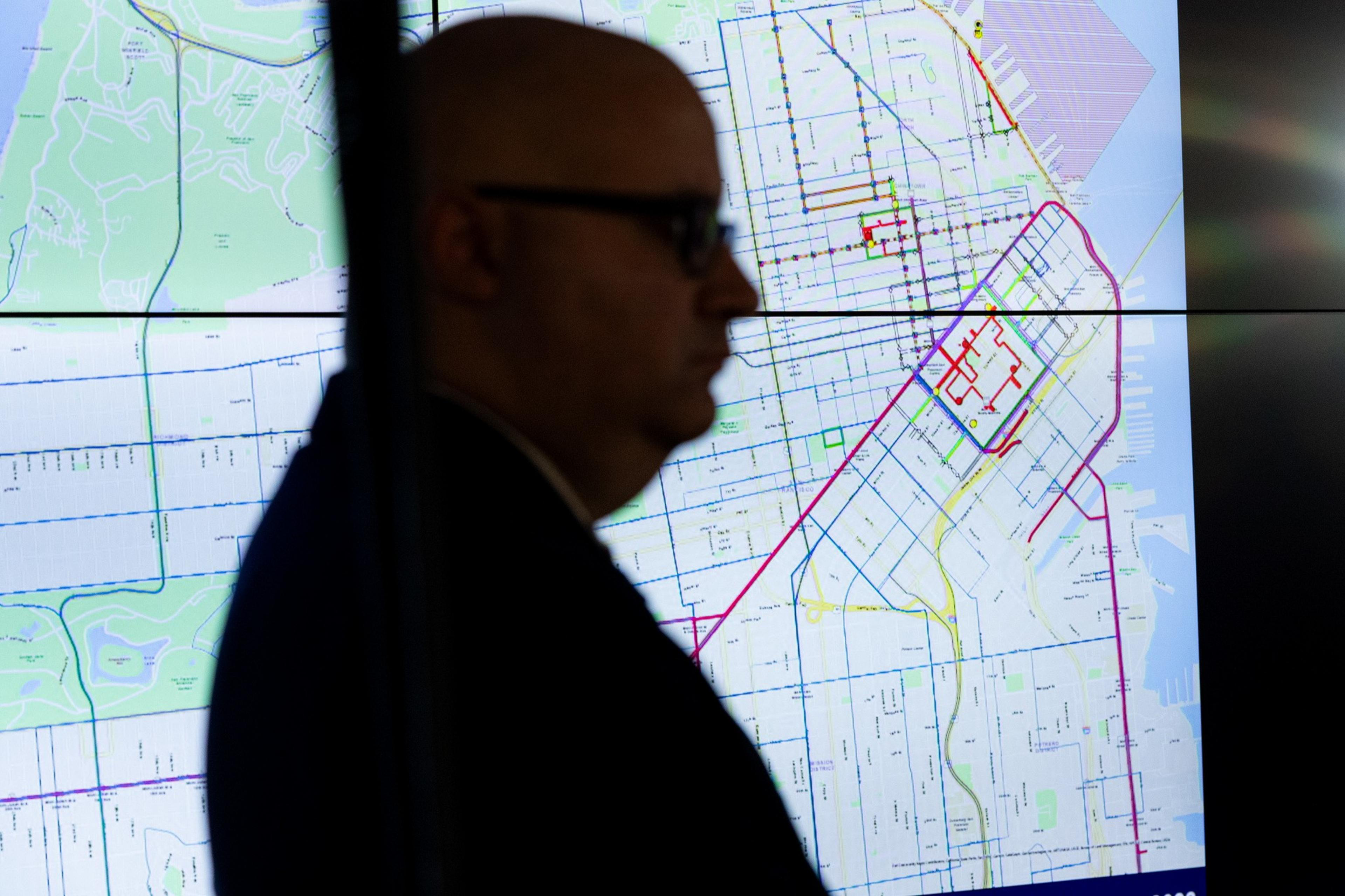 A man with glasses is silhouetted against a brightly lit, detailed city map displayed on a large screen behind him.