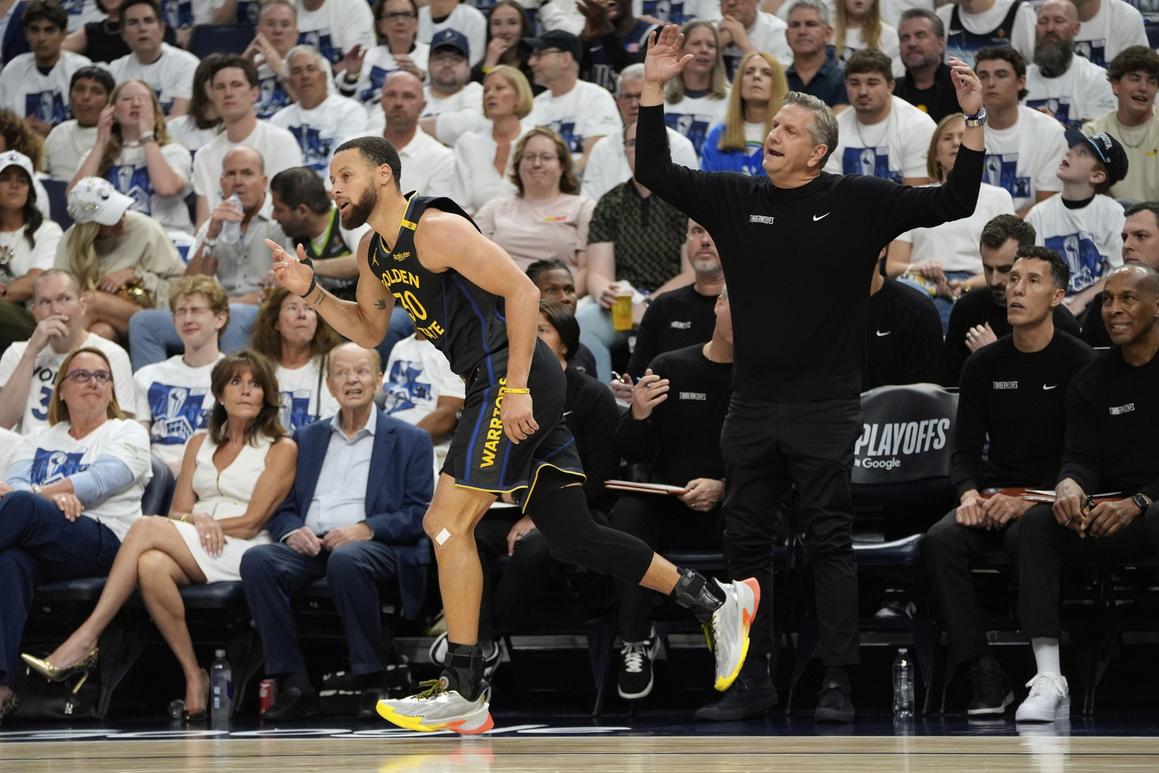 A basketball player in a Warriors uniform is running on the court. Nearby, a coach on the sideline gestures, while fans in white shirts watch intently.