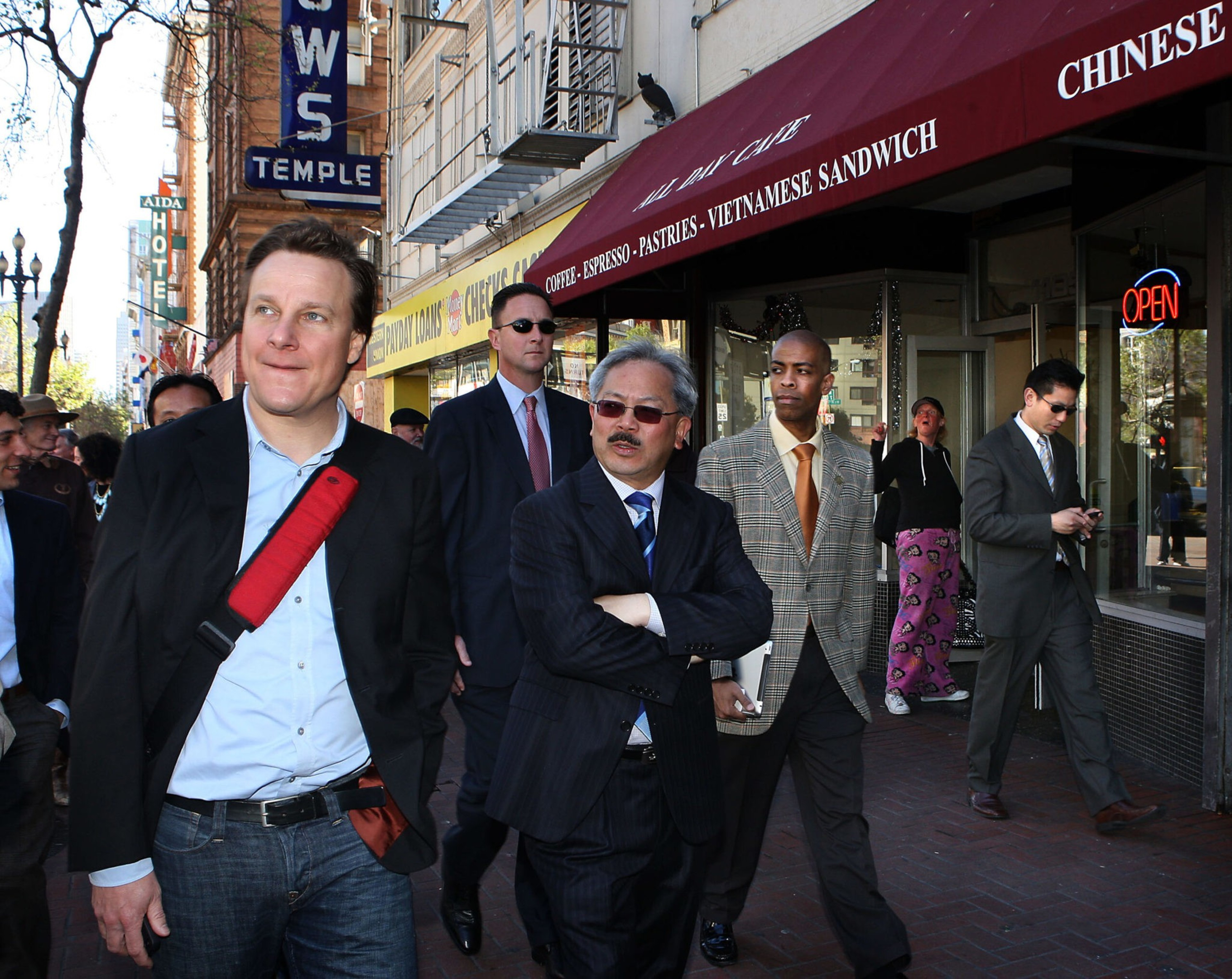 A group of men in suits and casual clothes walk past a cafe with a red awning advertising coffee and Vietnamese sandwiches.