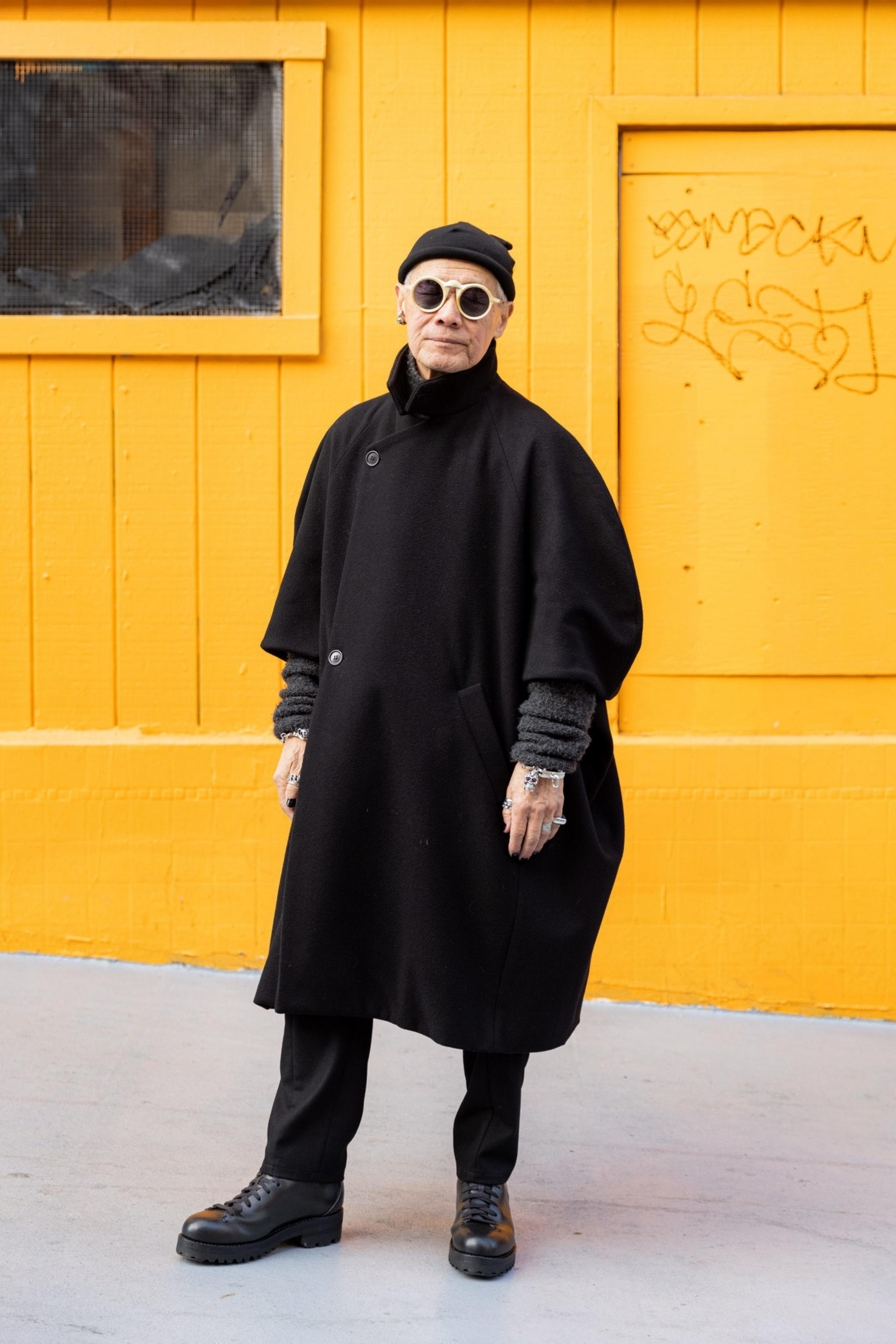 An elderly person wears a black oversized coat, beanie, round sunglasses, and boots, standing against a bright yellow wall with graffiti.